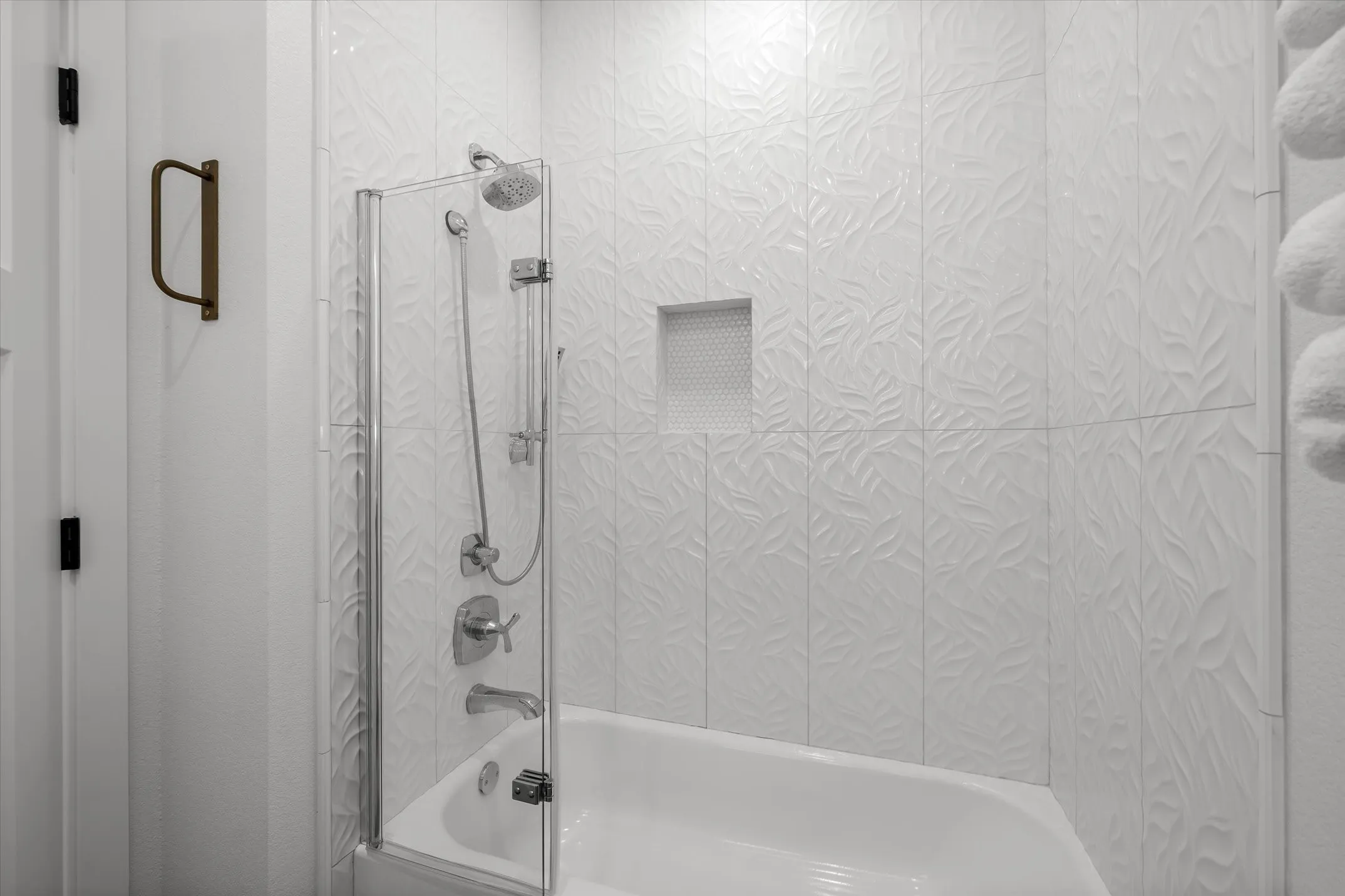 Bathtub with integrated shower, chrome fixtures, glass shower screen, and textured tile surround with recessed niche