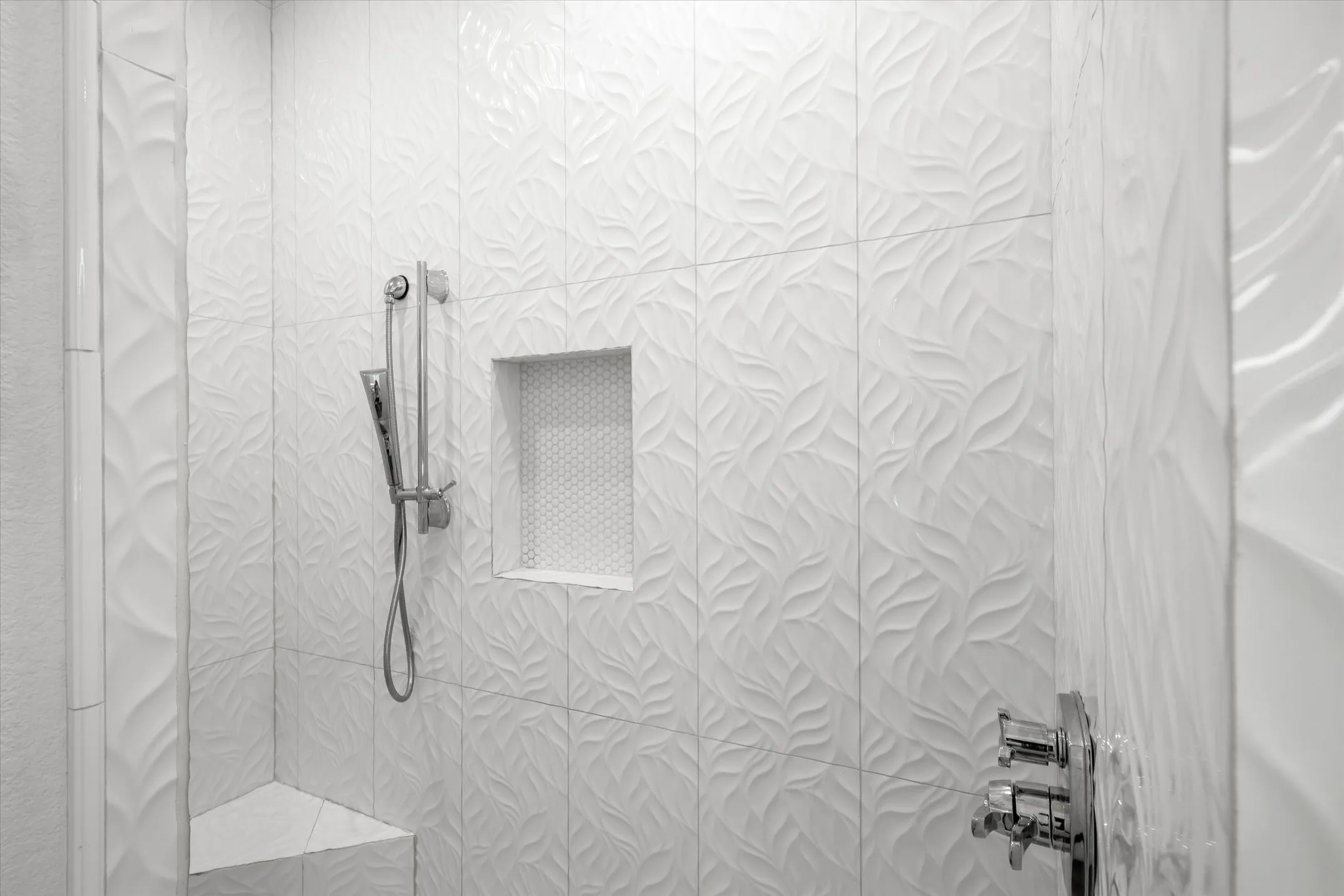 Textured white tile shower surround with a built-in niche, integrated shower bench, handheld shower fixture, and polished chrome controls