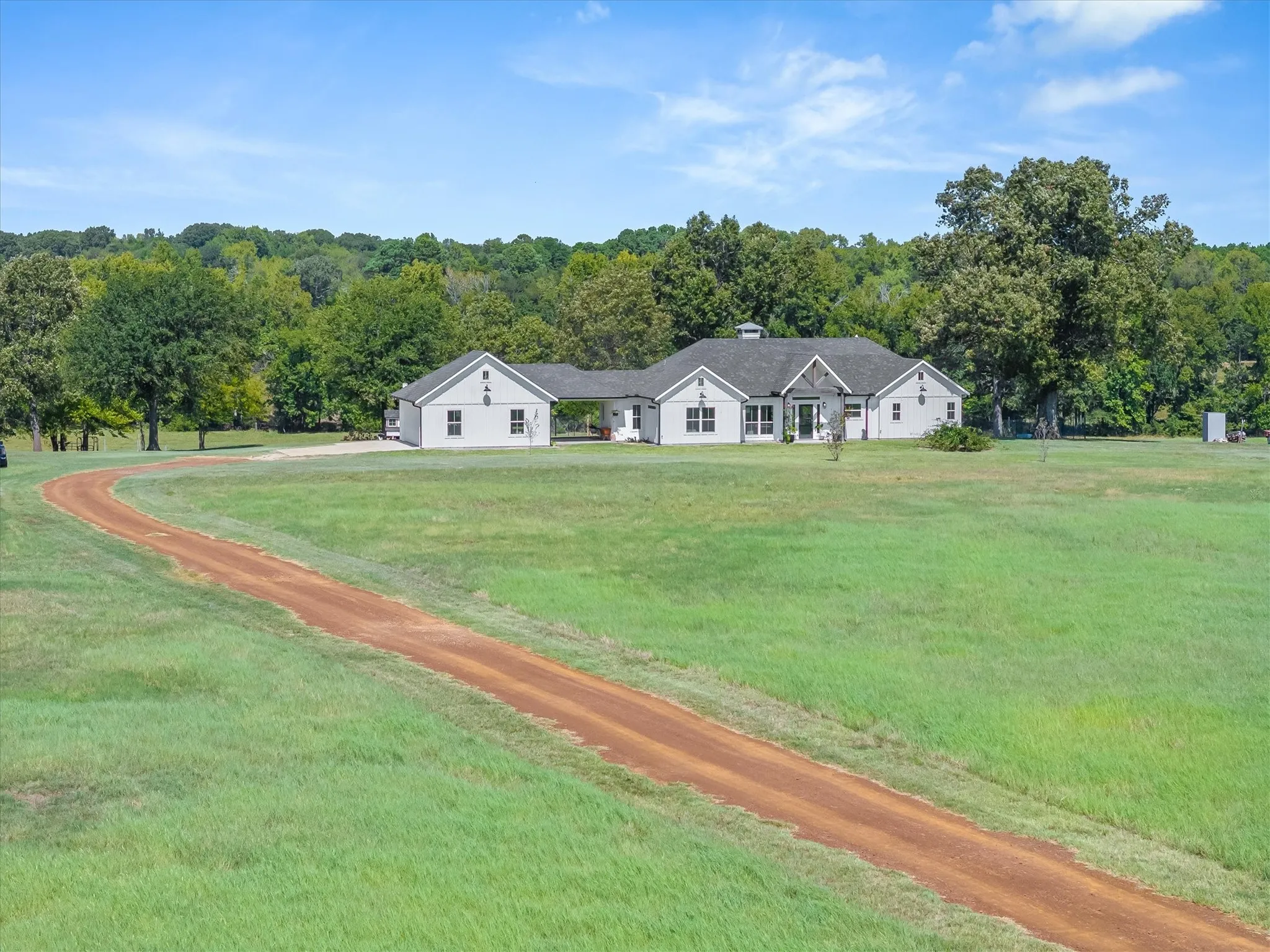 Expansive green lawn with a long, winding gravel driveway, leading to a single-story residence with a light-colored exterior and dark roof