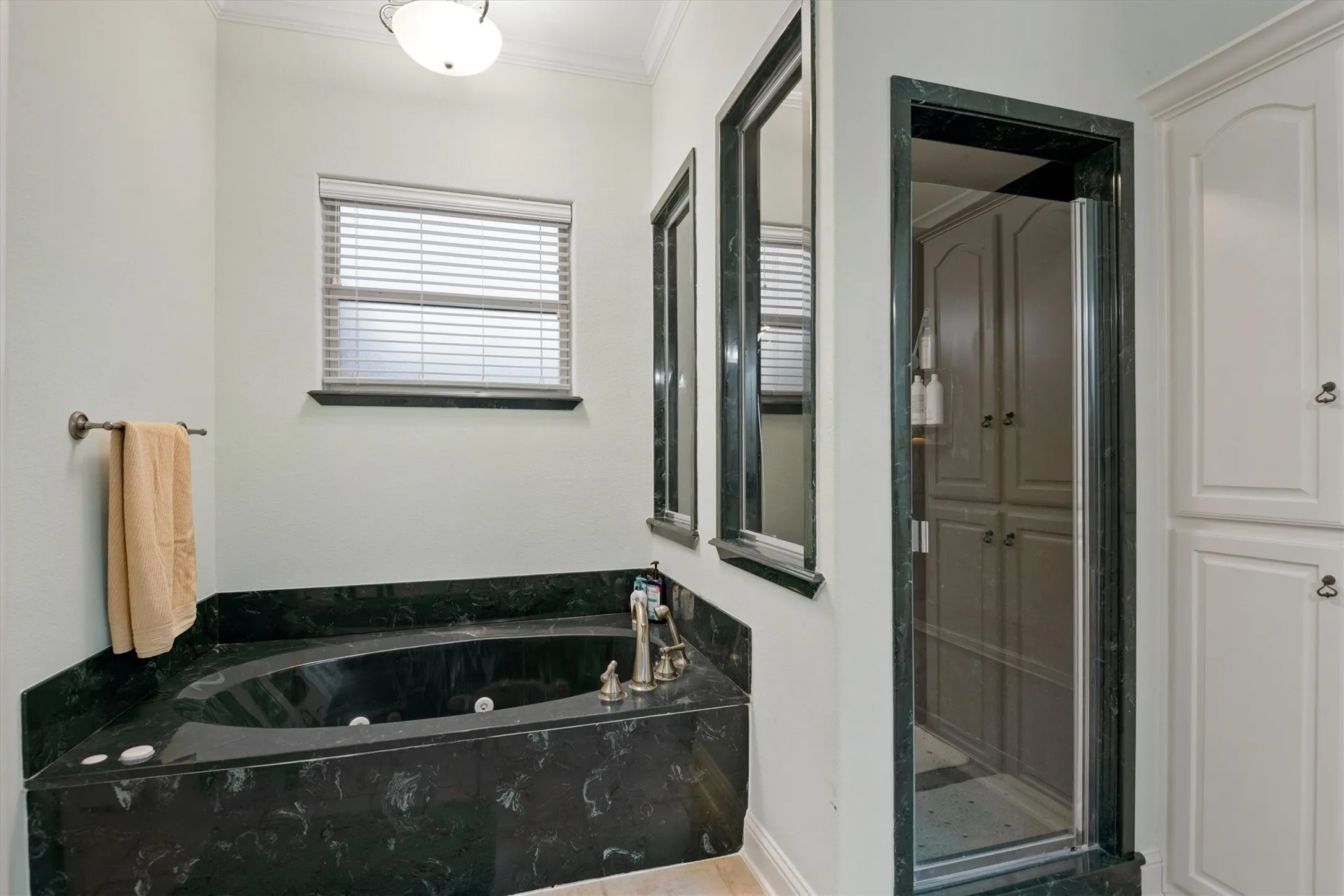 Built-in corner tub with dark stone surround, walk-in shower with glass enclosure, white paneled cabinetry, and window with blinds