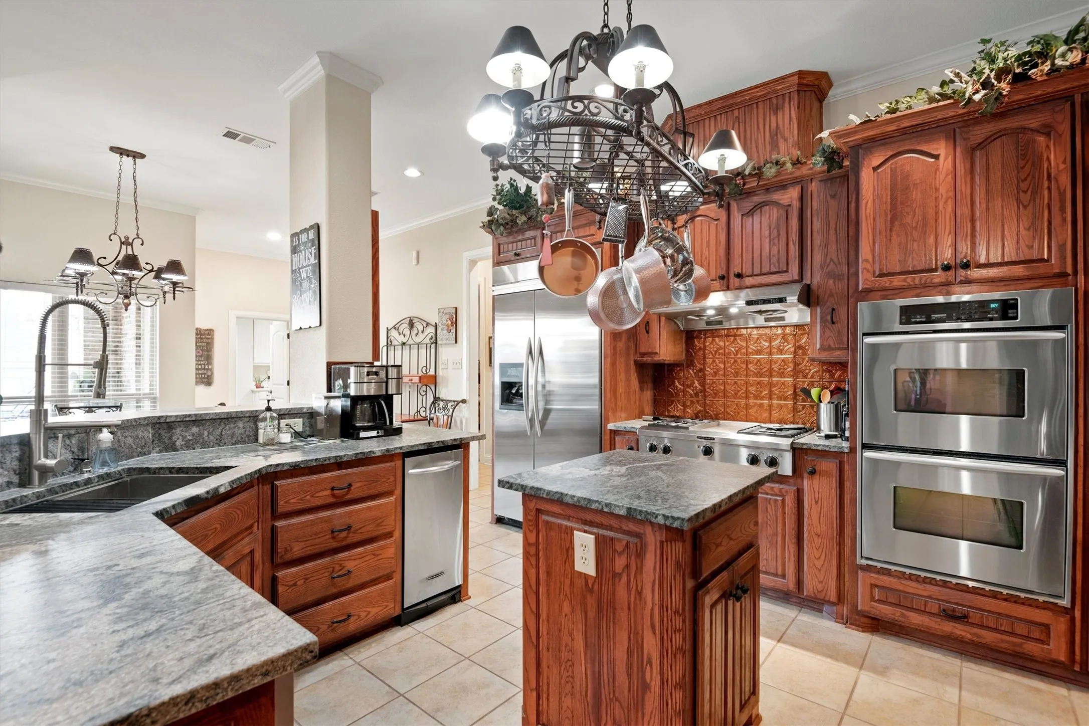 Kitchen featuring natural wood cabinetry, light-toned granite countertops, stainless steel appliances, a center island, and a pot rack chandelier