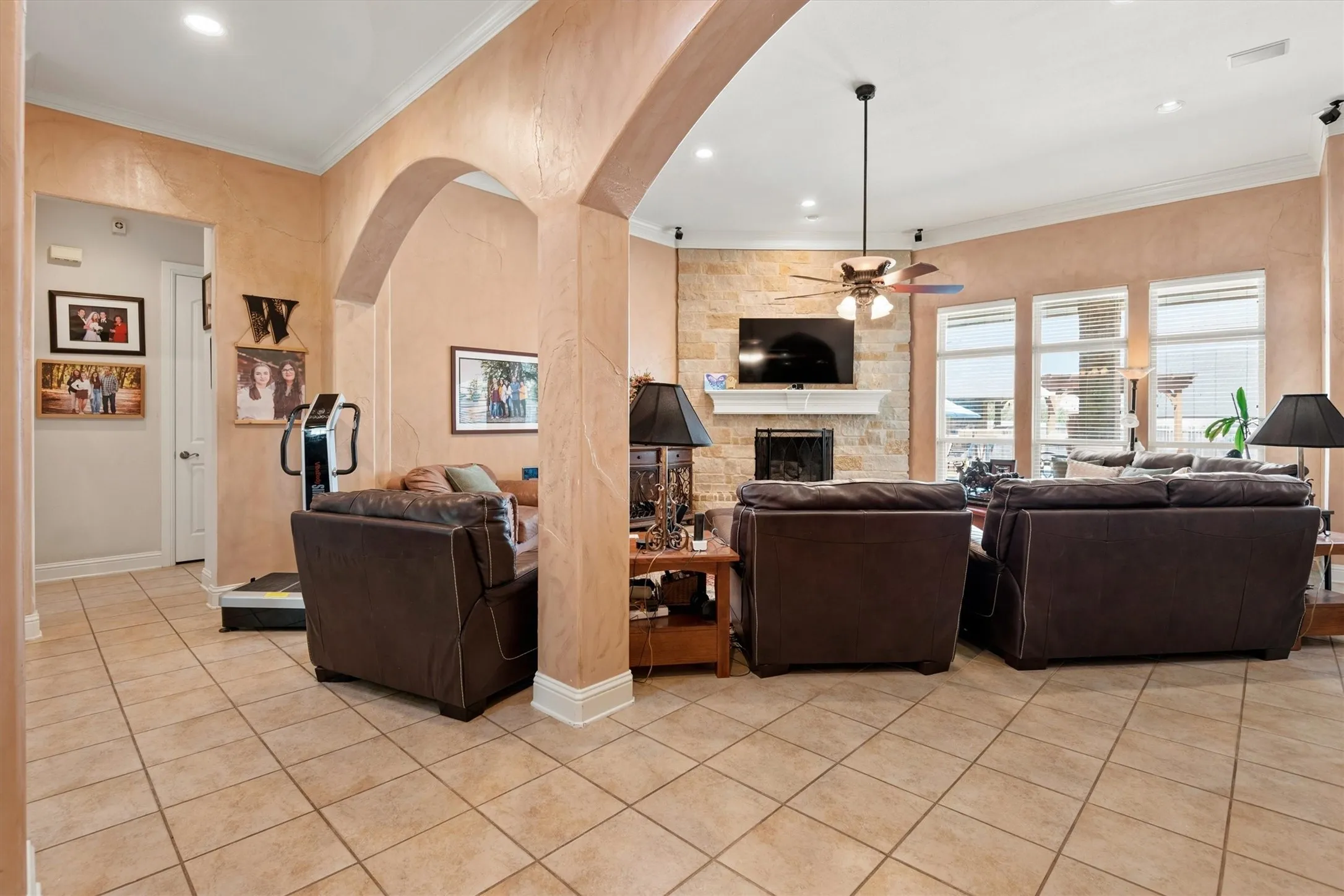 Arched entryway with crown molding, recessed lighting, tile flooring, stone fireplace with mantel, and large windows with blinds