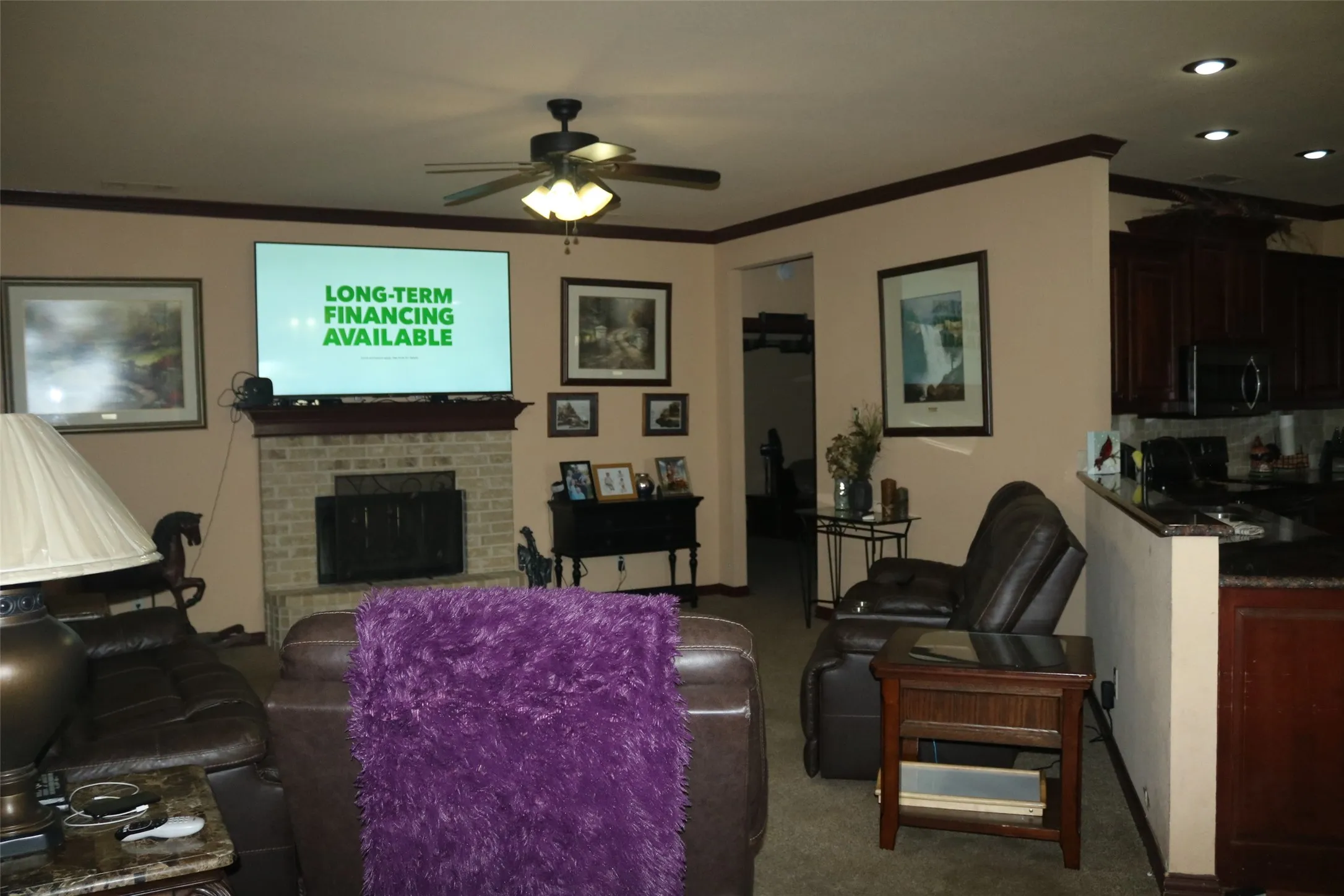 Brick-front fireplace with a wood mantel, ceiling fan with integrated lighting, recessed lighting, wood-finish cabinetry, and dark countertops