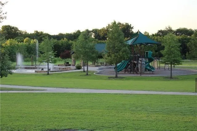 Expansive green space featuring a playground with a shade structure, a water fountain feature, mature trees, and paved walking paths