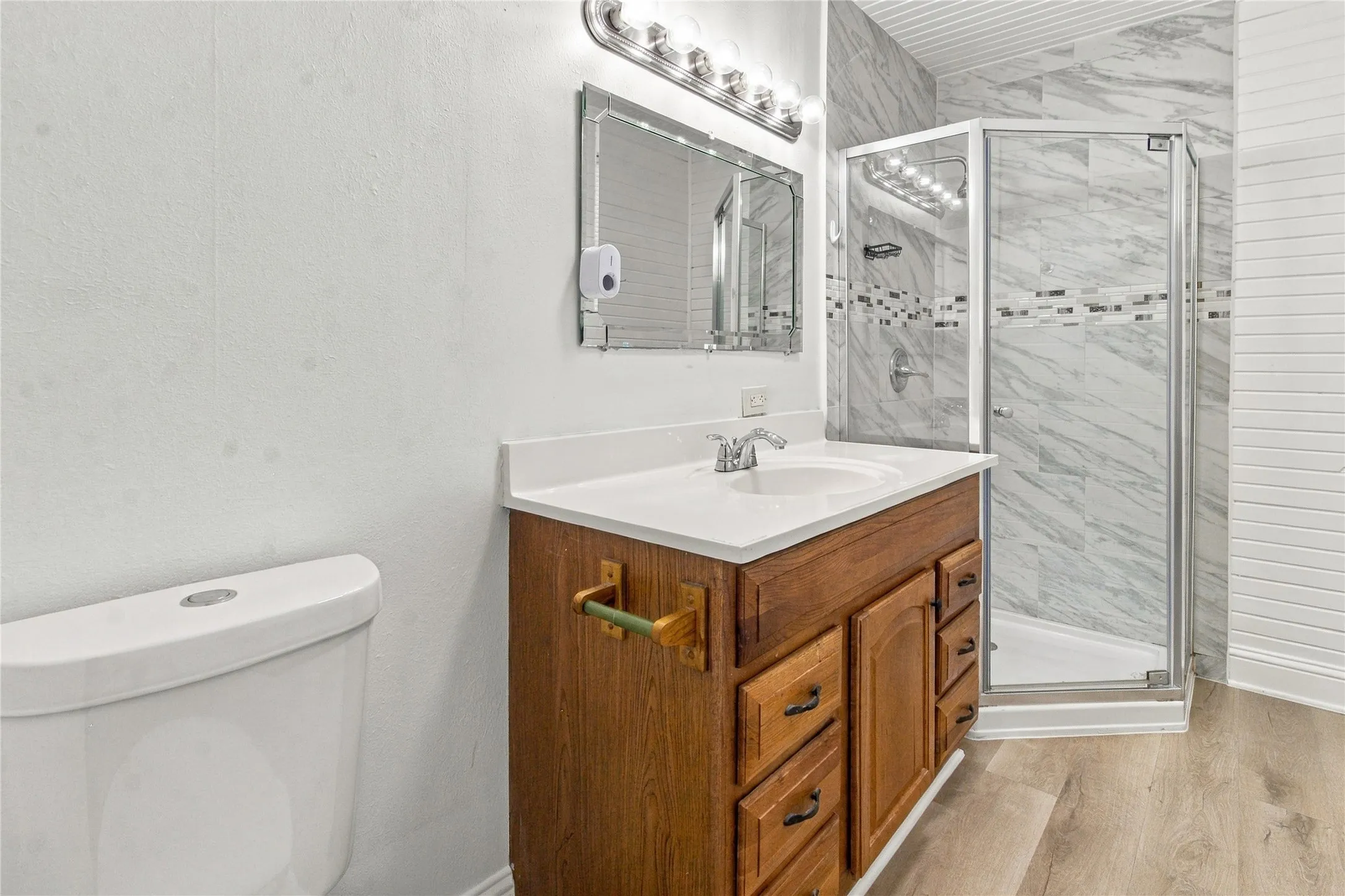 Wood-finish flooring, single vanity with a white countertop, chrome faucet, and a frameless glass shower enclosure with marble-style tile