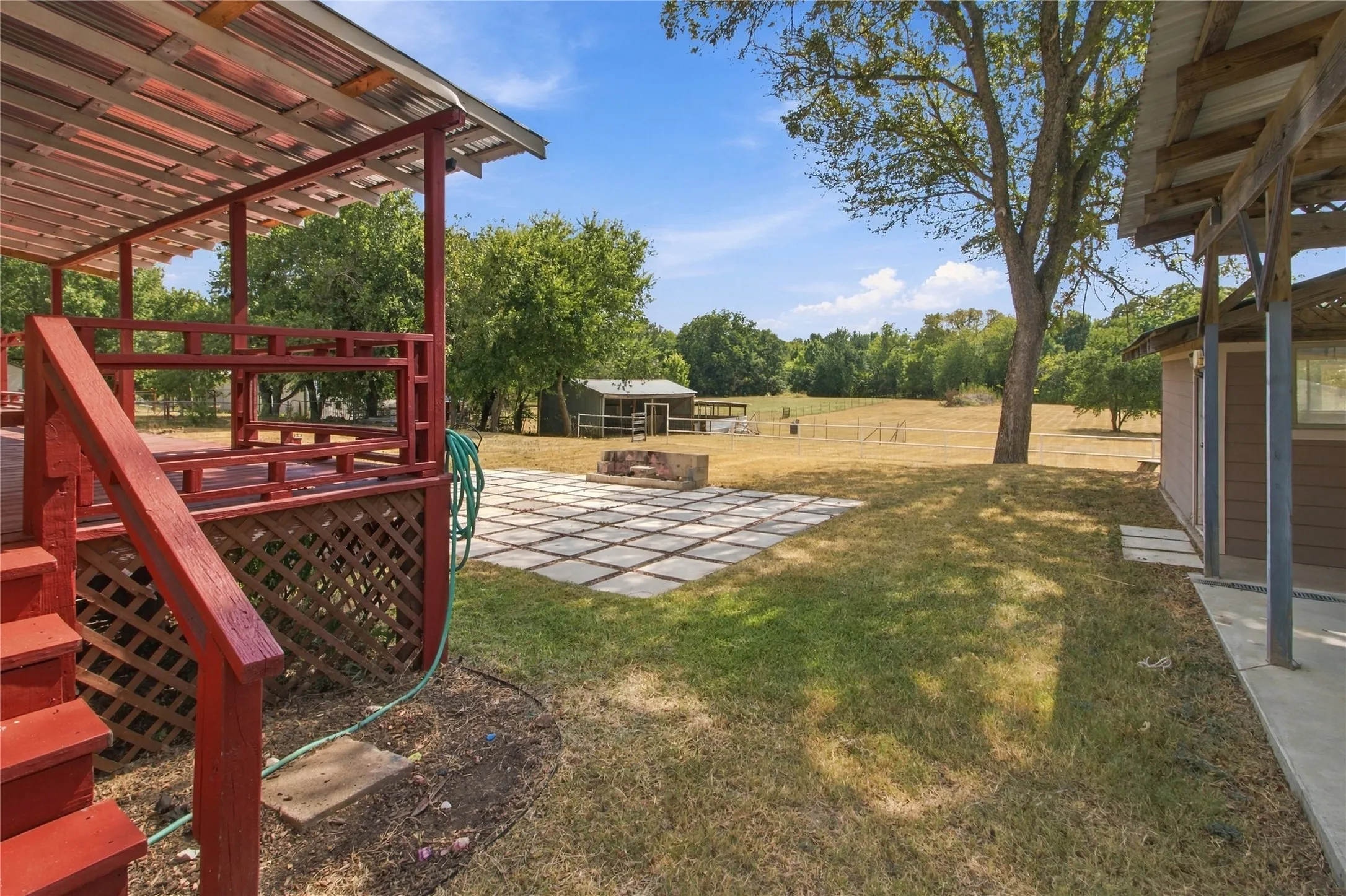 Covered outdoor deck with red-painted railings and lattice accents, expansive grass area, concrete paver patio, separate detached structure, and mature trees