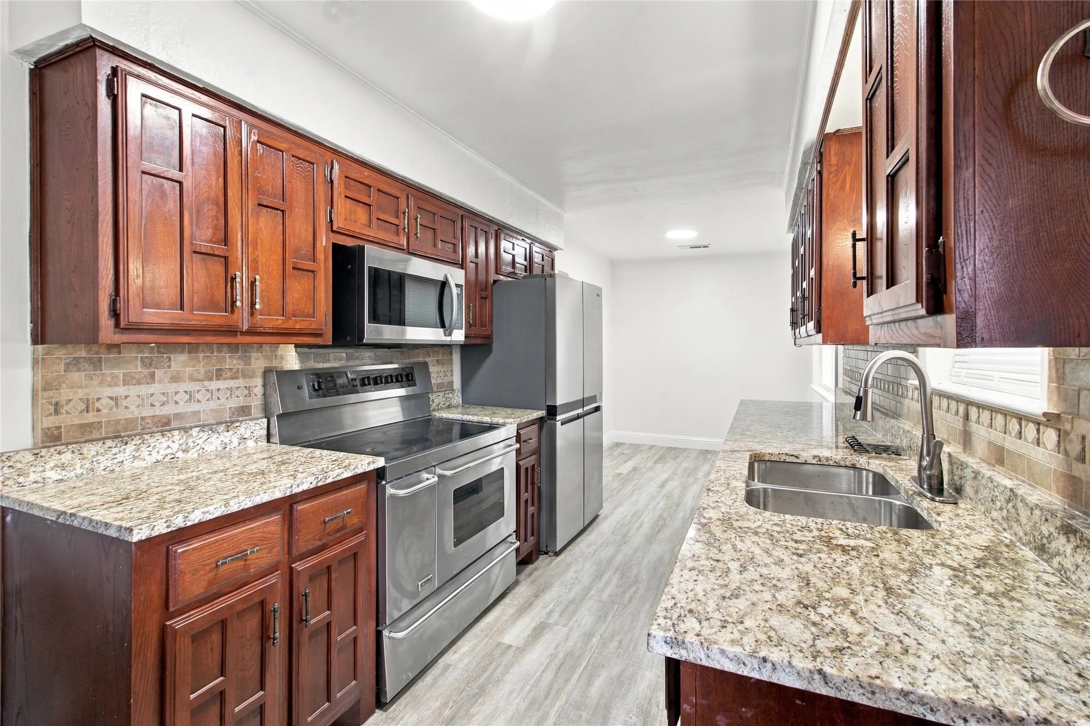 Kitchen featuring wood-finish cabinetry, granite-style countertops, stainless steel appliances, a tile backsplash, and light-tone wood-finish flooring