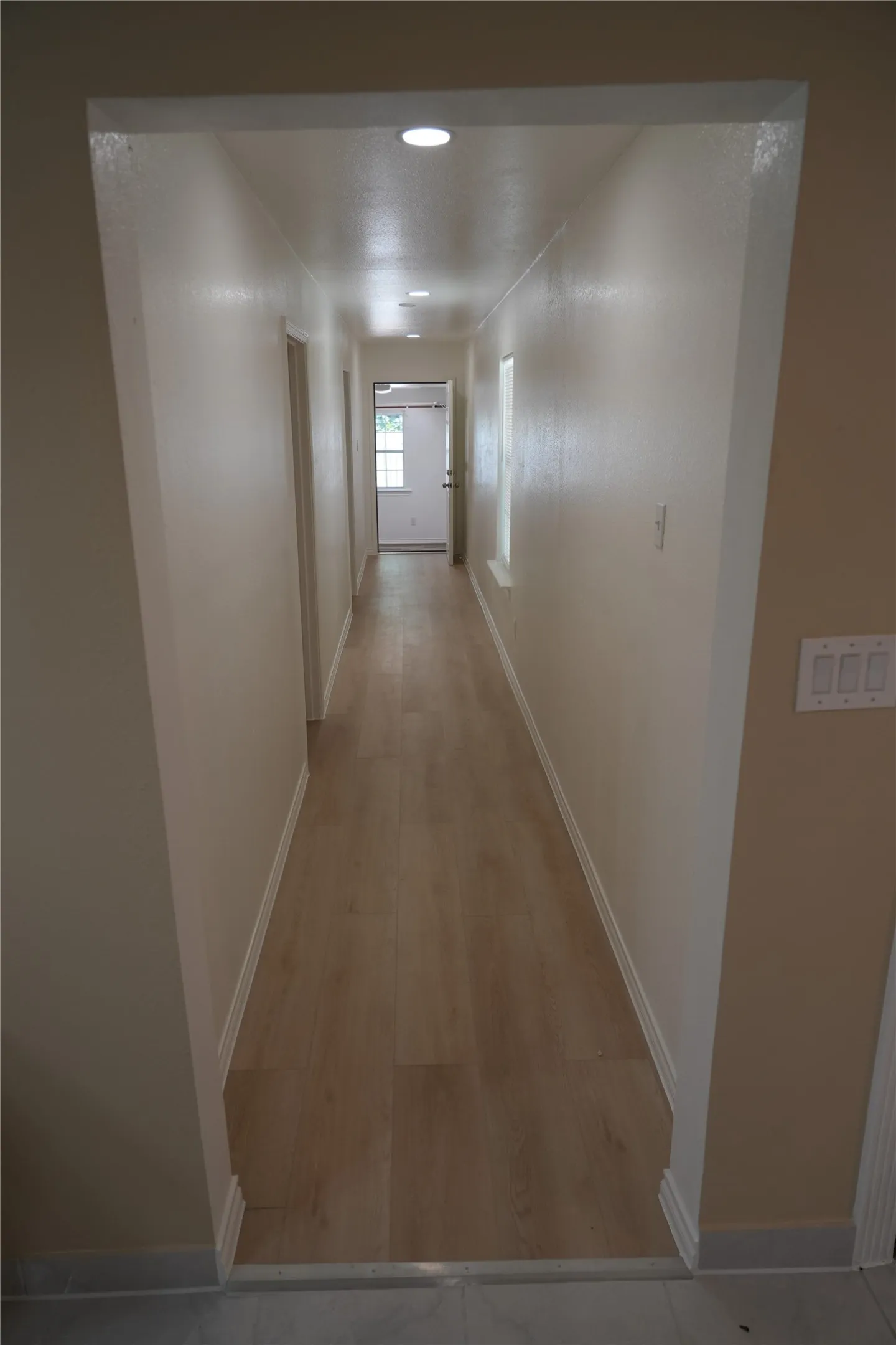 Recessed lighting illuminates a long hallway with wood-finish flooring and light-toned walls