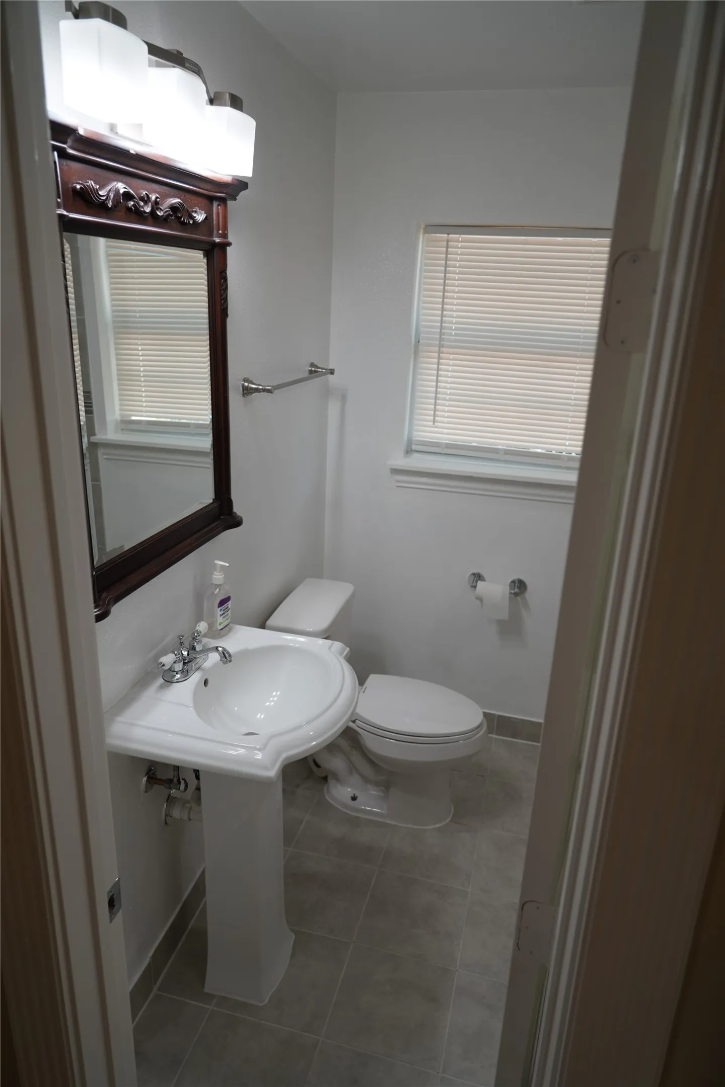 Pedestal sink with chrome faucet, a toilet, a window with blinds, a wall-mounted towel bar, and a framed mirror with an overhead light fixture