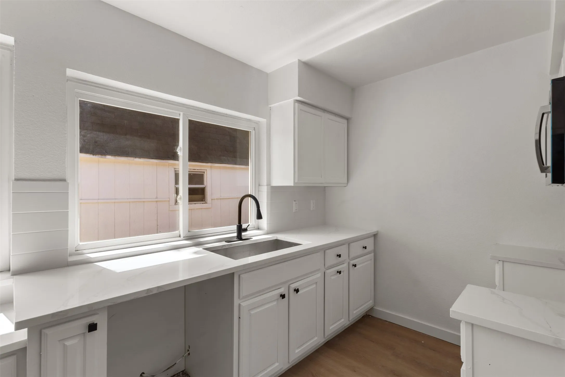 White shaker-style cabinetry with black hardware, an undermount stainless steel sink, a black gooseneck faucet, light-colored countertops, and wood-finish flooring