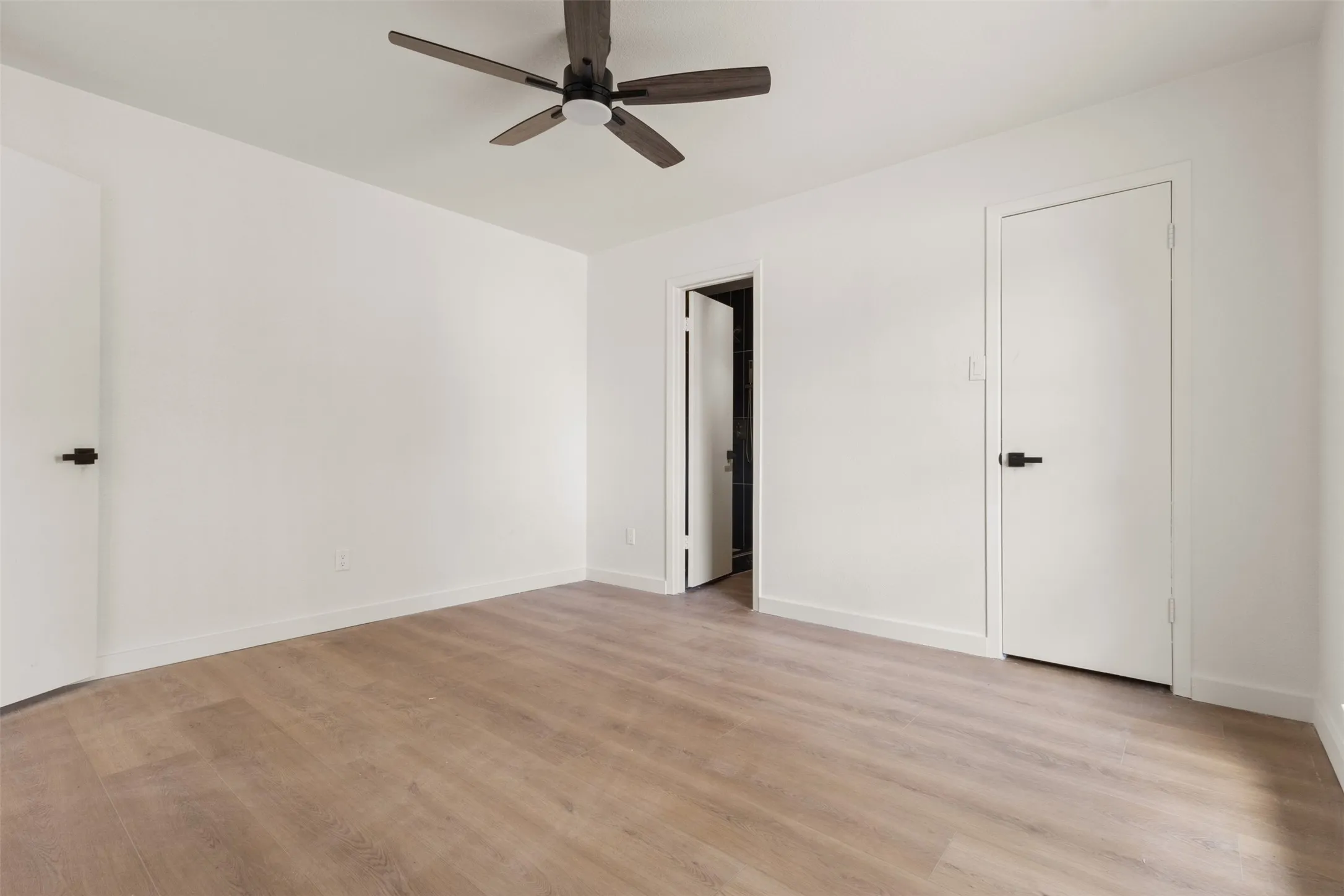 Wood-finish flooring, white walls, dark ceiling fan, and white interior doors with matte black hardware