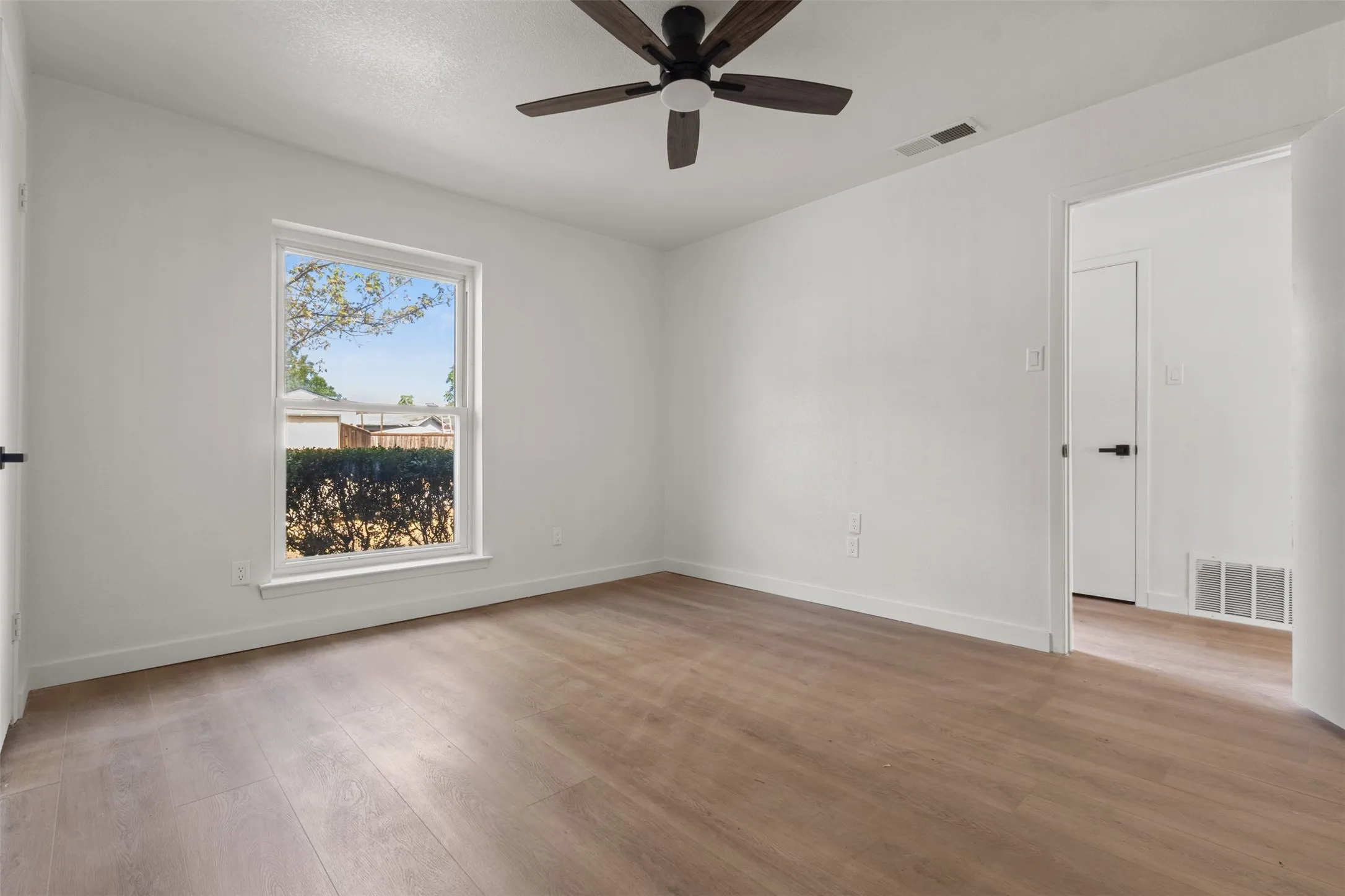 Wood-finish flooring, white painted walls, a large window, a dark ceiling fan, and a white door with dark hardware