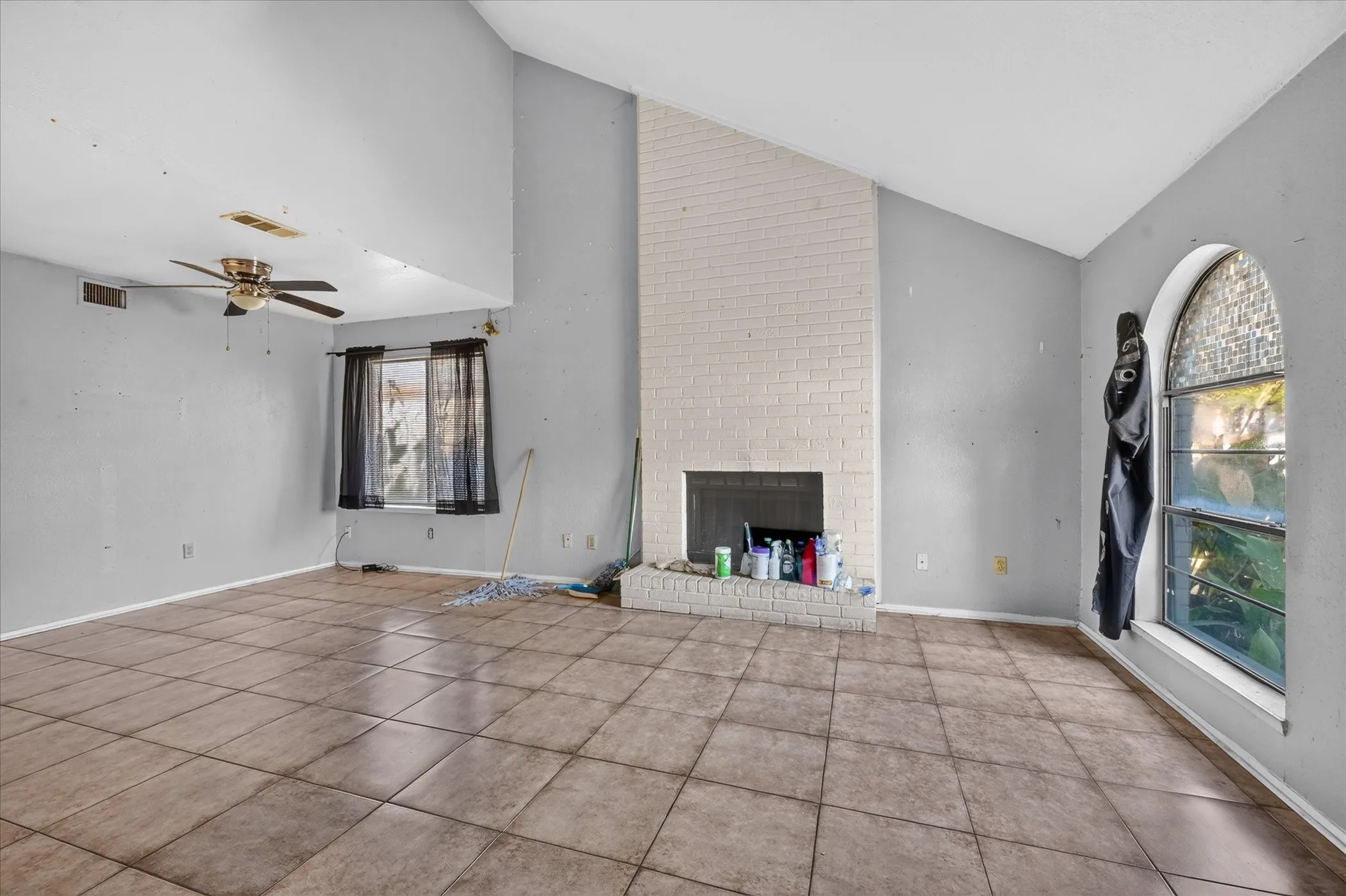 Vaulted ceiling, brick-surround fireplace, tile flooring, arched window, and ceiling fan with light fixture