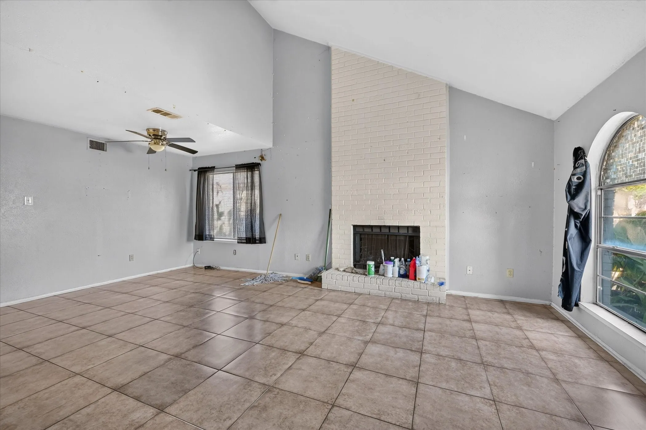 Vaulted ceiling with a ceiling fan, tiled flooring, painted brick fireplace, and an arched window