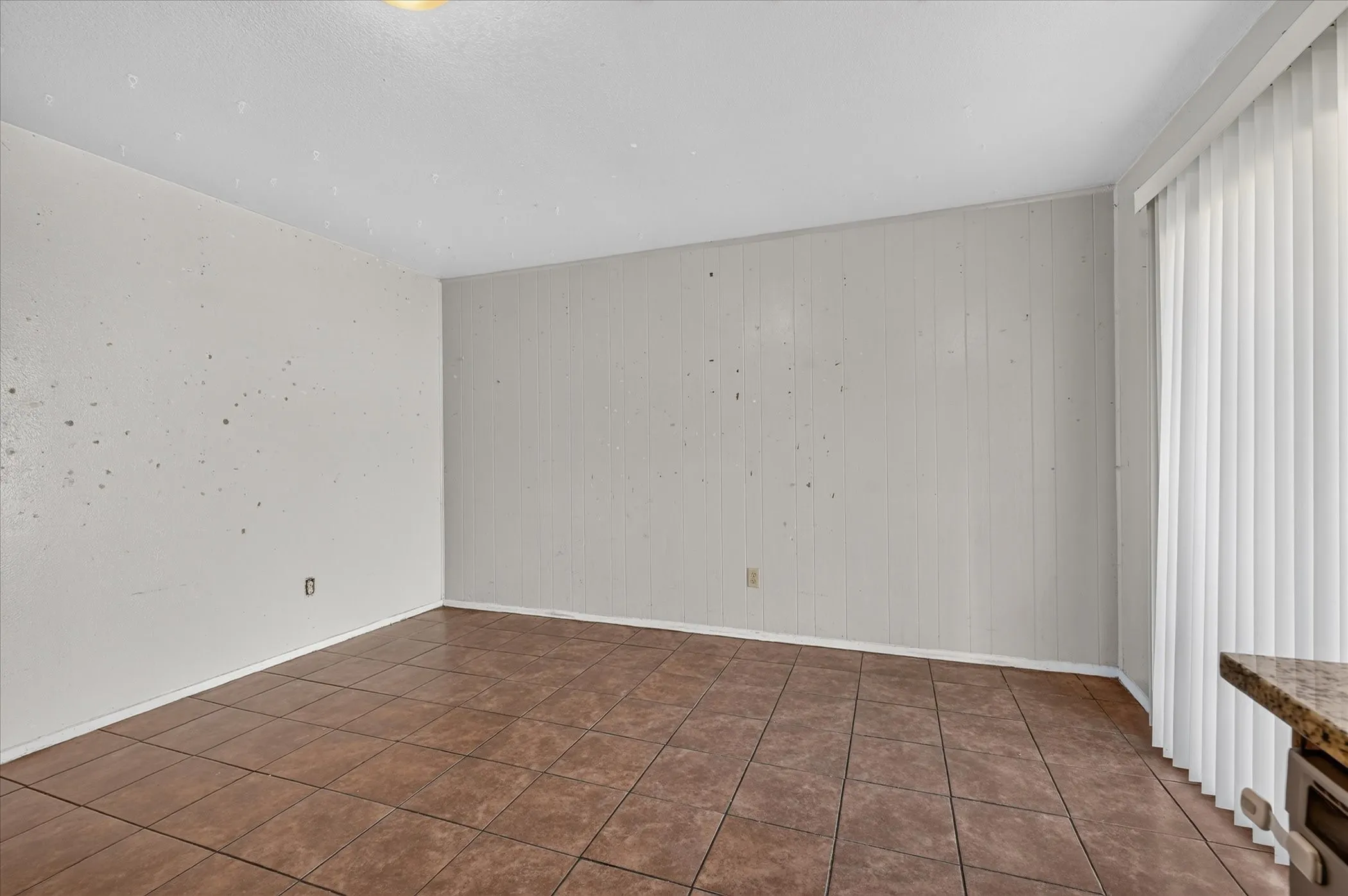 Tile flooring, vertical paneling accent wall, single flush-mount ceiling light, vertical blinds, and a built-in counter surface with stone-finish