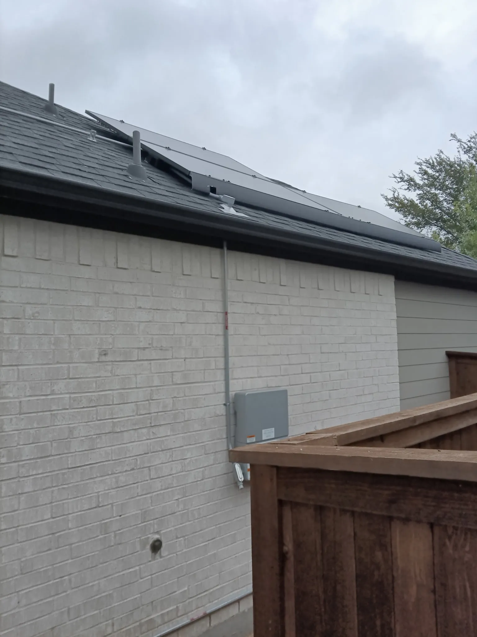 Roof-mounted solar panels, dark shingle roof, white brick exterior, wood fence, and grey siding