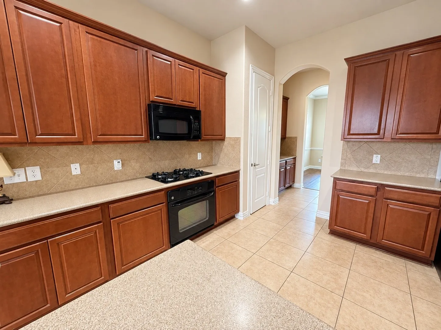 Cherry wood-finish cabinetry, built-in black appliances, tan tile flooring, and light-colored countertops