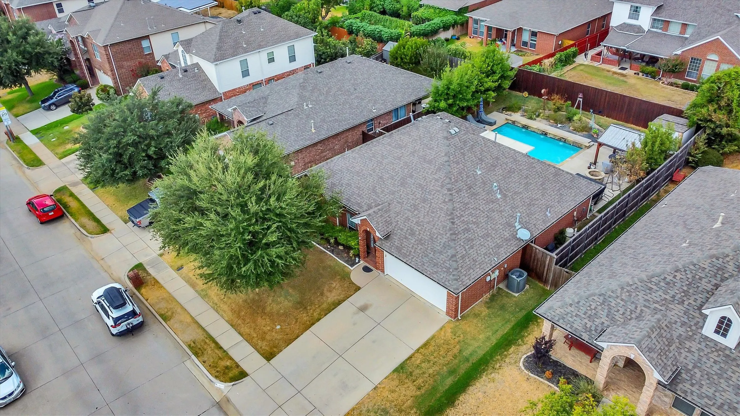 Brick exterior with an attached garage, concrete driveway, front yard trees, fenced backyard, and an in-ground pool