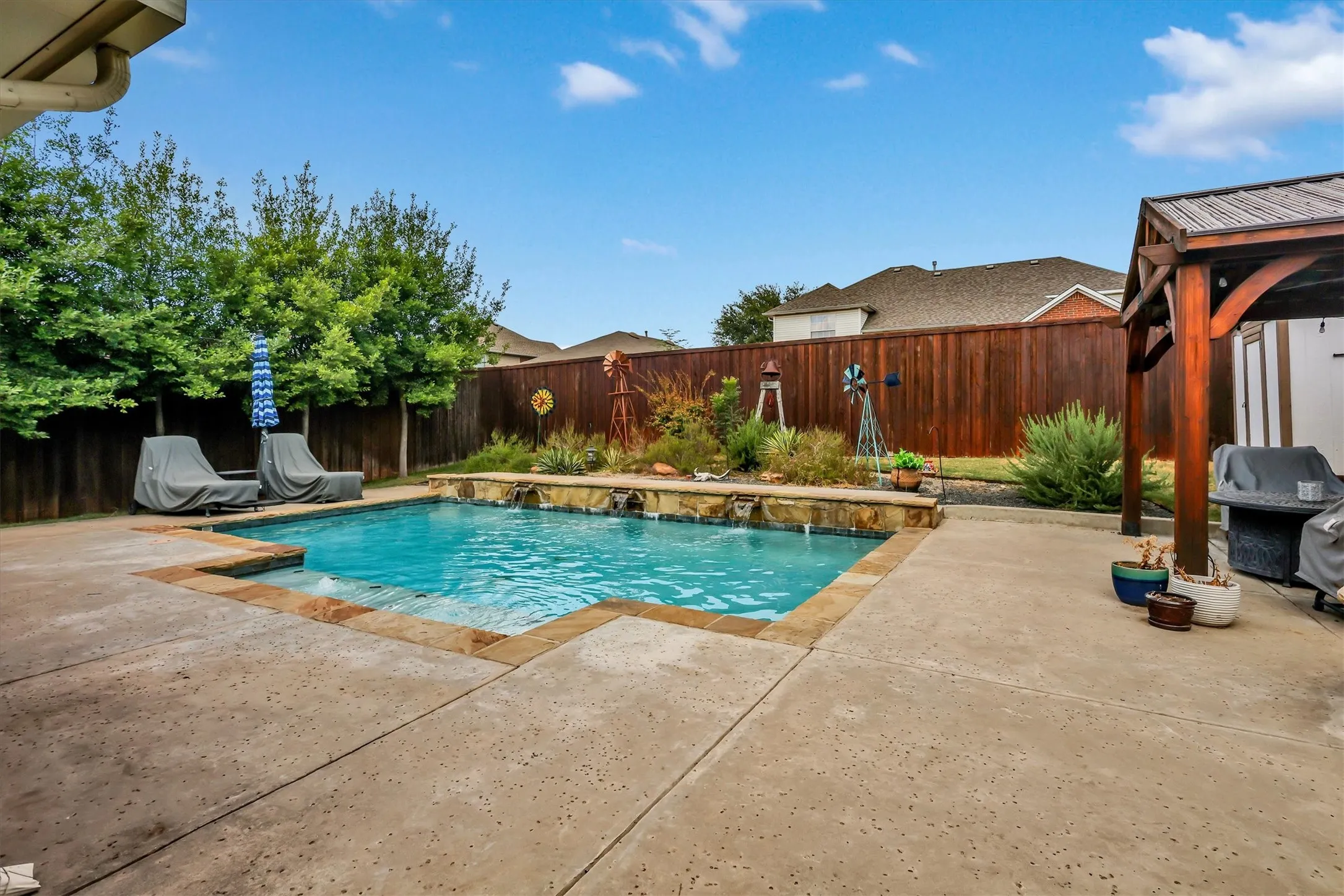 Expansive concrete patio surrounding a built-in swimming pool with water features, a wood-framed pergola, and a privacy fence