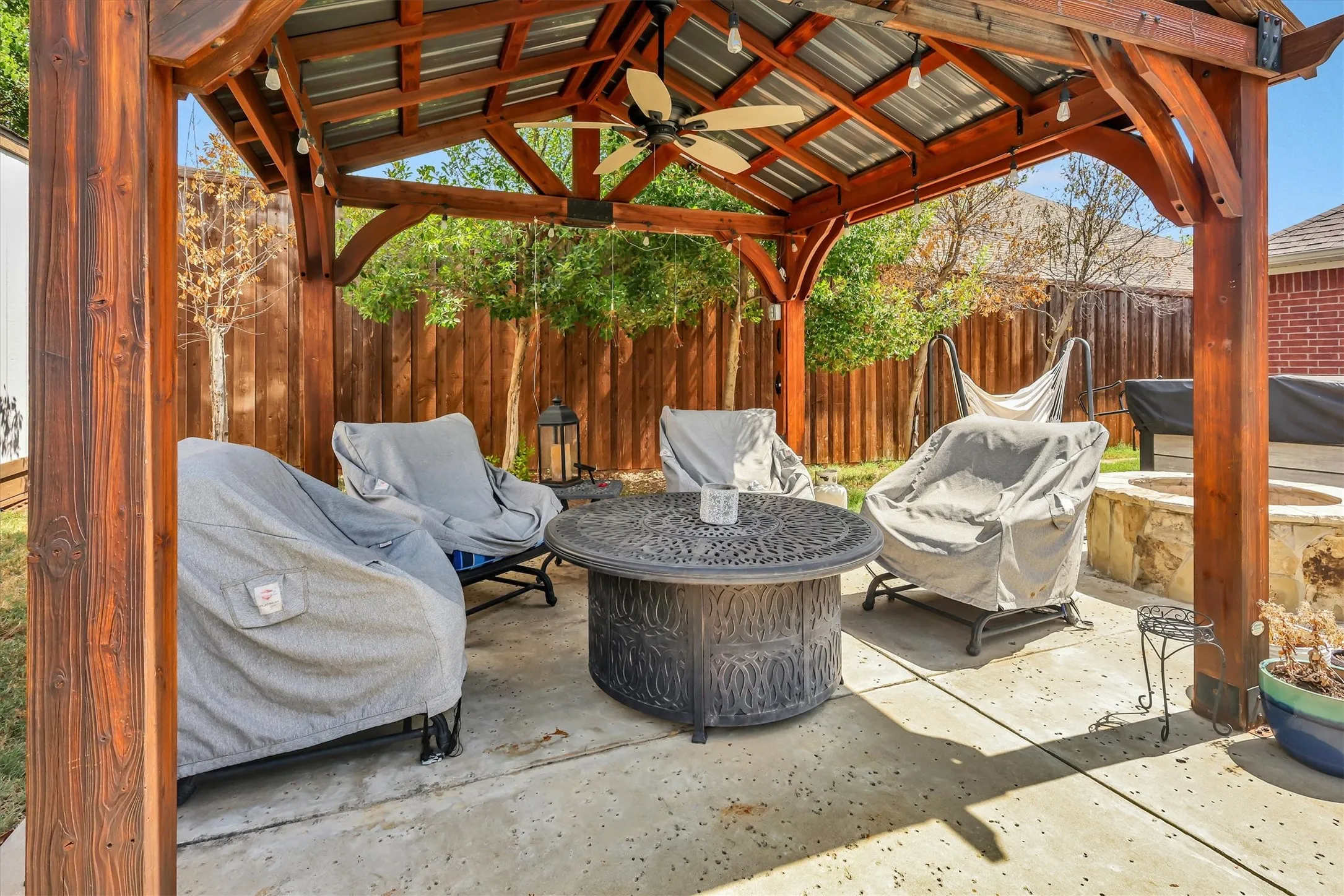 Patio featuring a wood pergola with a metal roof, a ceiling fan, string lighting, and a built-in fire pit with stone surround