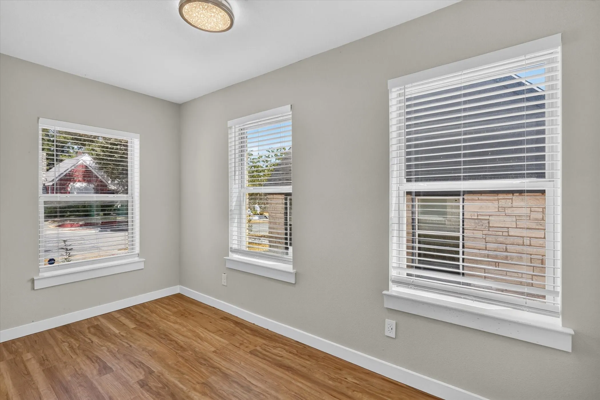 Room featuring three windows with blinds, wood-finish flooring, light gray walls, and a ceiling-mounted light fixture