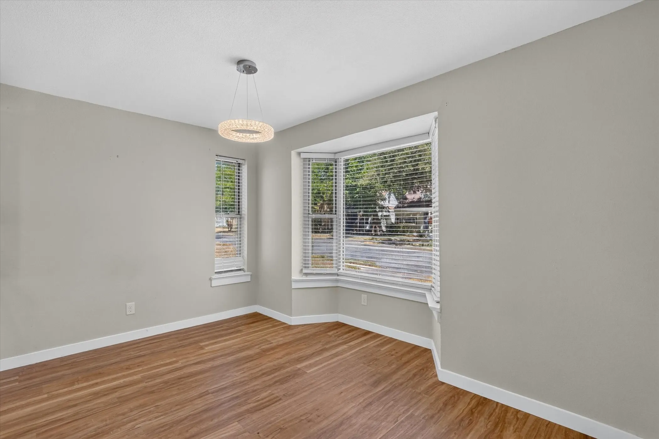 Hardwood-finish flooring, bay window with blinds, modern chandelier, and light gray wall paint
