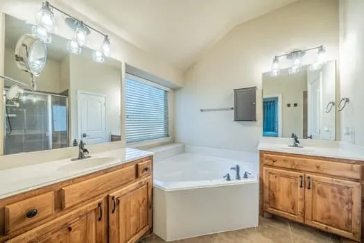 Dual wood-finish vanities with countertop sinks, a built-in corner soaking tub, a separate glass-enclosed shower, and a vaulted ceiling