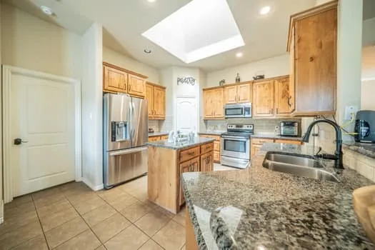 Kitchen with natural wood cabinetry, stainless steel appliances, a center island with a sink, granite-style countertops, and a skylight