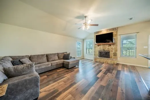 Vaulted ceiling featuring a ceiling fan, stone-clad fireplace, wood-finish flooring, and multiple windows