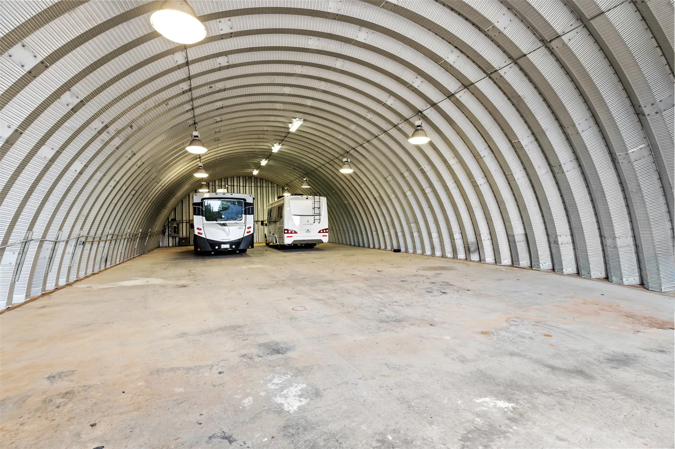 Arched corrugated metal structure with concrete flooring and overhead lighting