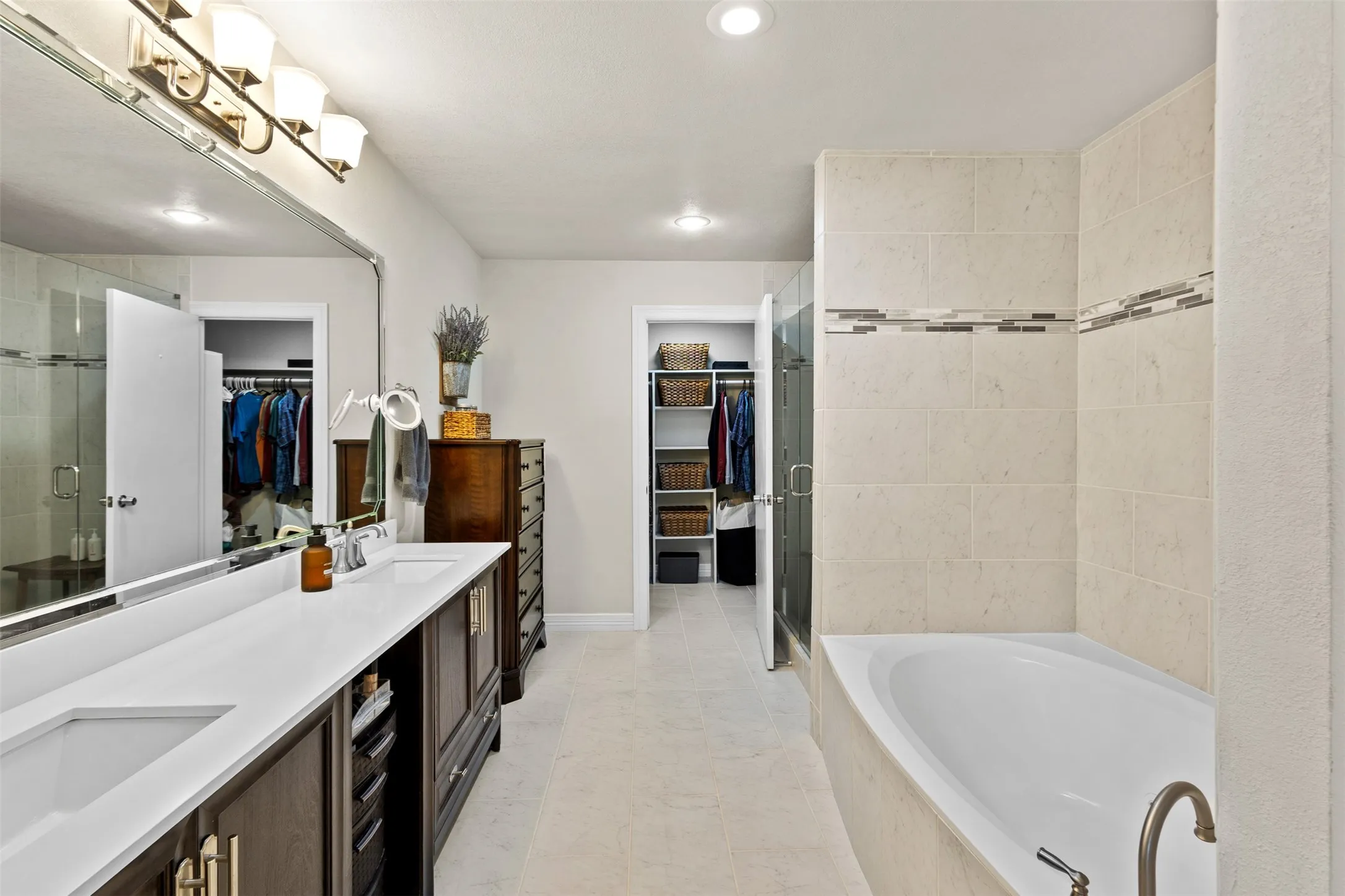 Bathroom featuring a dual vanity with white countertops, expansive wall mirror, built-in bathtub, and a glass-enclosed shower