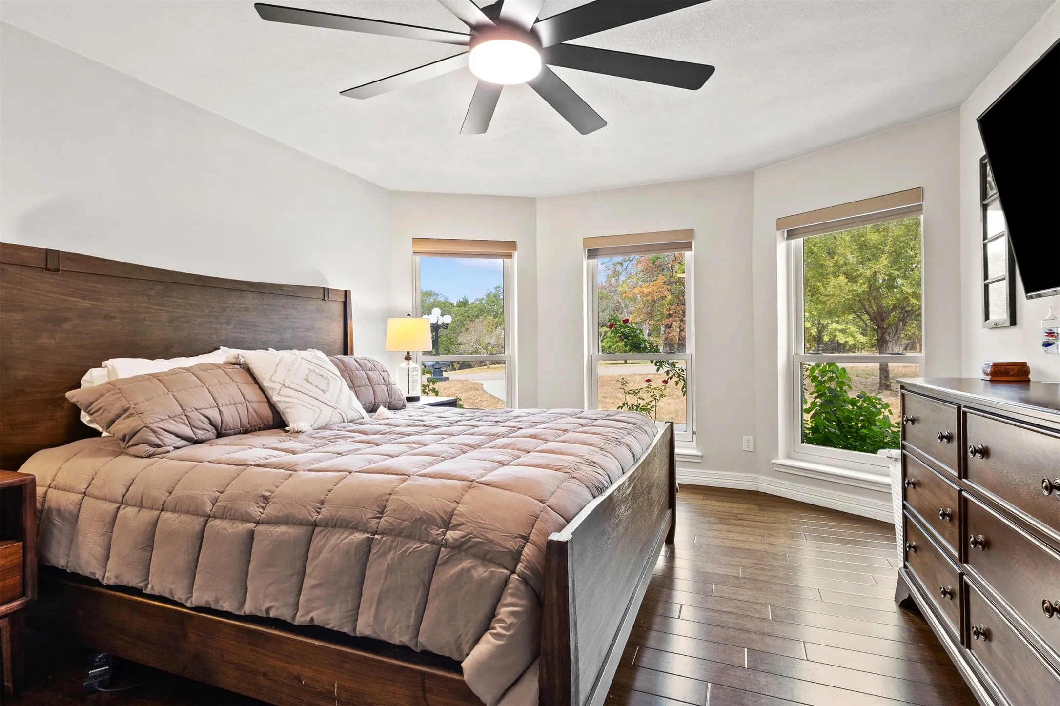 Primary bedroom suite featuring a wood-finish platform bed, abundant natural light from multiple glass sliding doors and windows, wide-plank wood flooring, ceiling fan with integrated lighting, and a built-in dresser with white cabinetry and marble-top surface