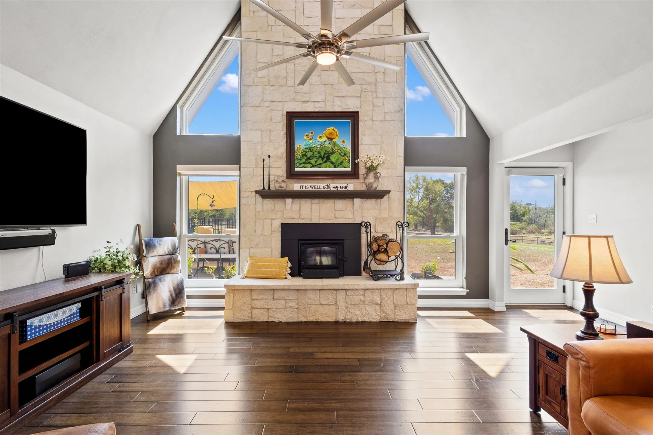 Vaulted ceiling, stone fireplace with hearth and mantel, wood-finish flooring, and multiple windows