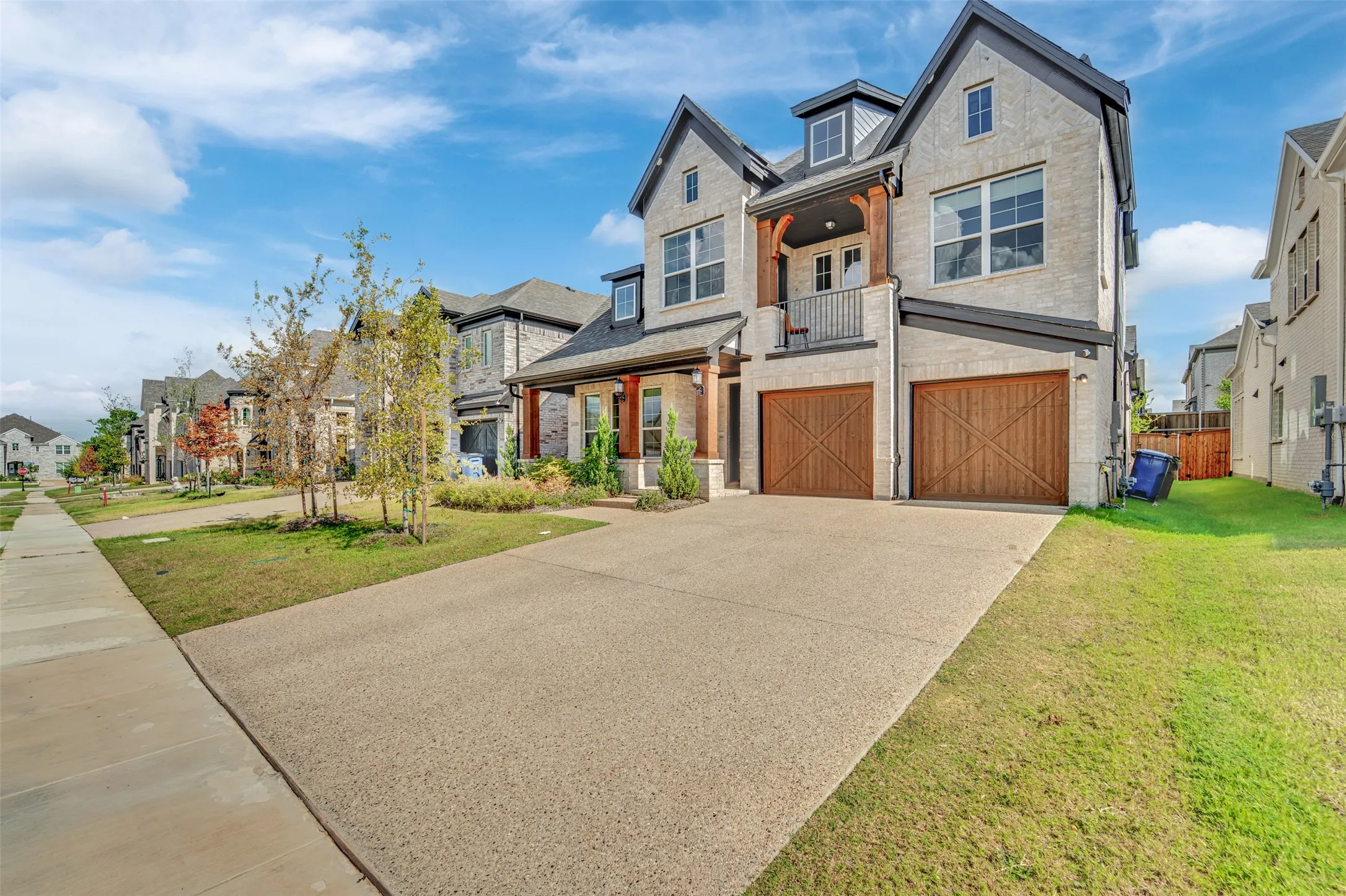 Stone exterior with dark trim accents, double garage with wood-finish doors, second-story balcony with iron railing, dormer windows, and a concrete driveway