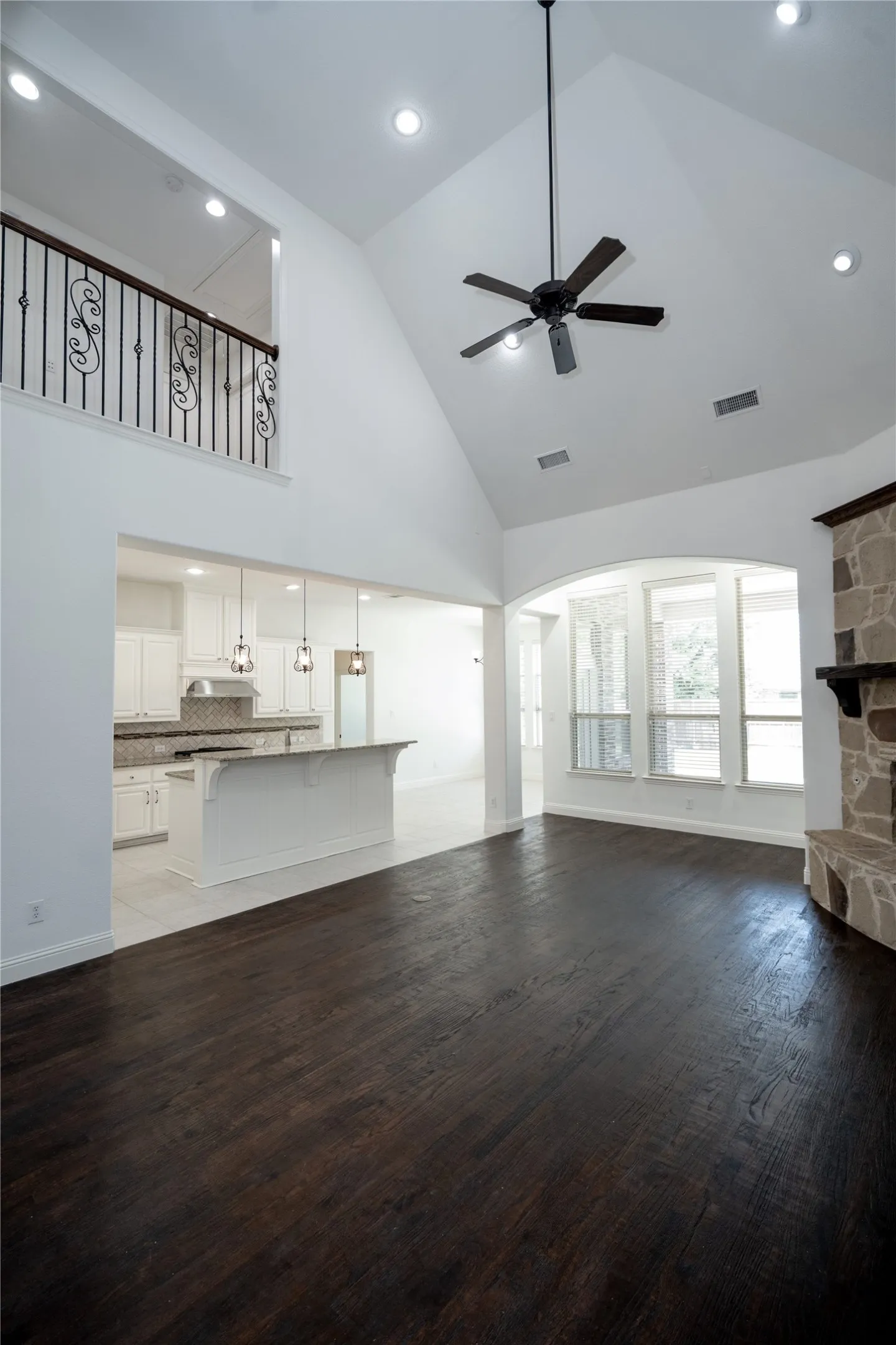 Vaulted ceiling with a ceiling fan, dark wood-finish flooring, stone fireplace, and an open-concept kitchen with a breakfast bar
