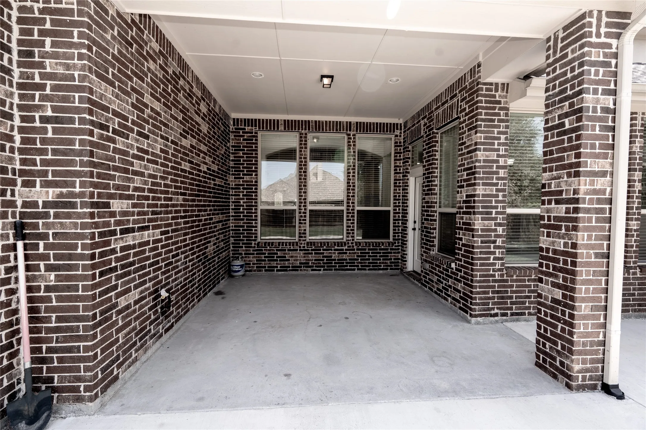 Covered concrete patio featuring dark brick walls, multiple windows, a white door, and a recessed ceiling light