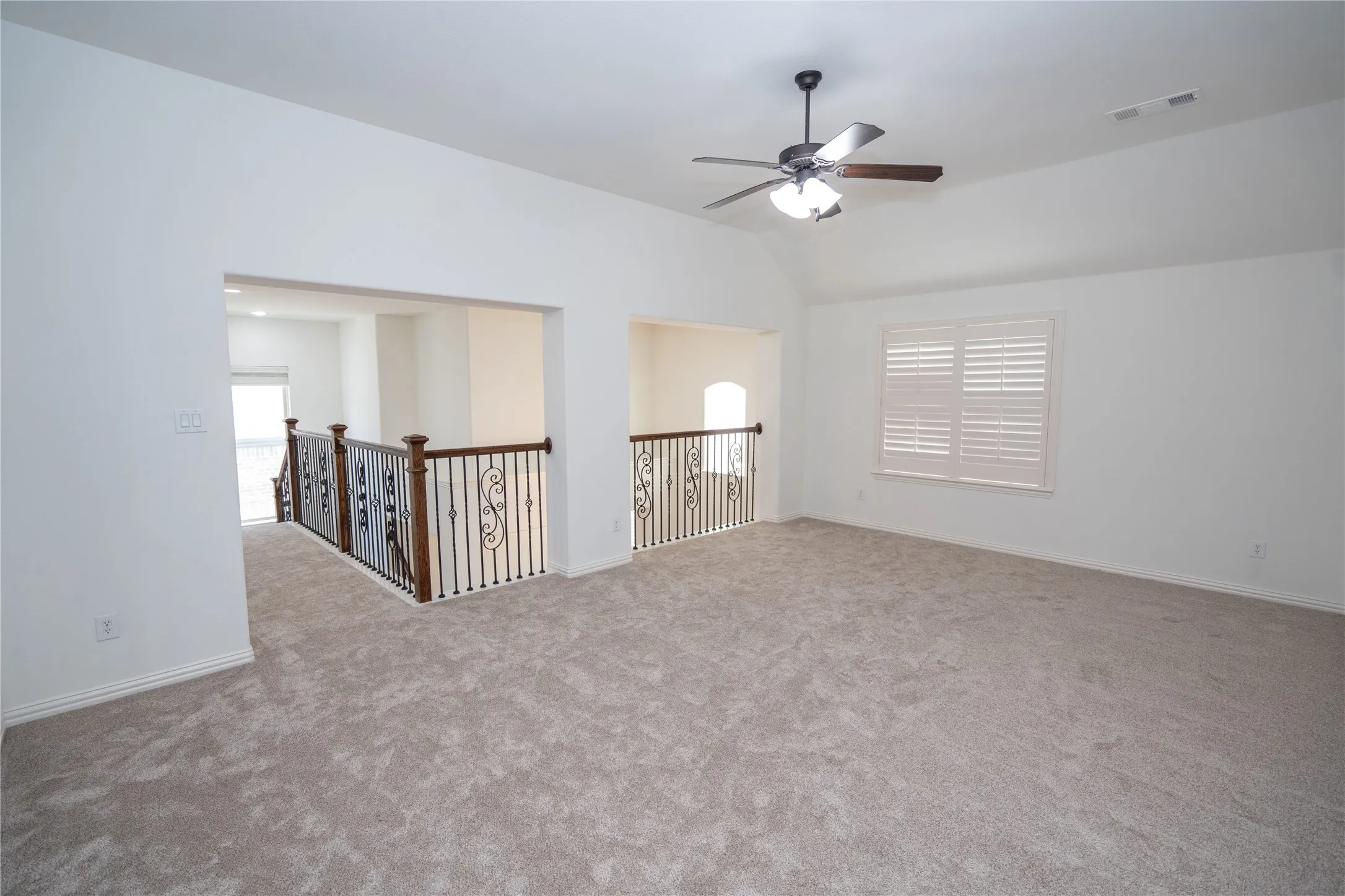 Carpeted loft space featuring a vaulted ceiling, ceiling fan with light, window with plantation shutters, and ornate metal railings