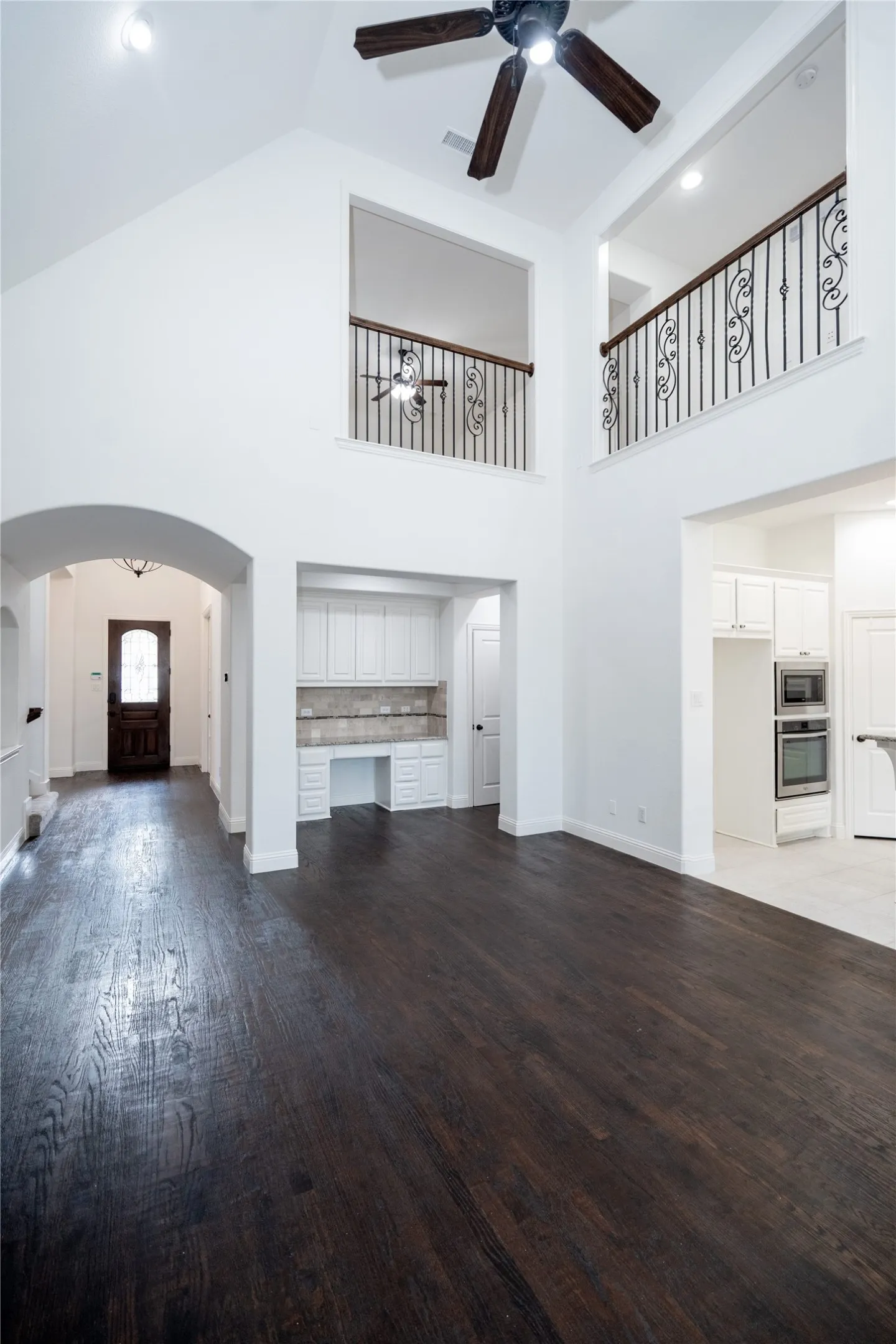 Soaring vaulted ceilings with a ceiling fan, dark wood-finish flooring, white built-in cabinetry with a desk area, and a wrought iron banister