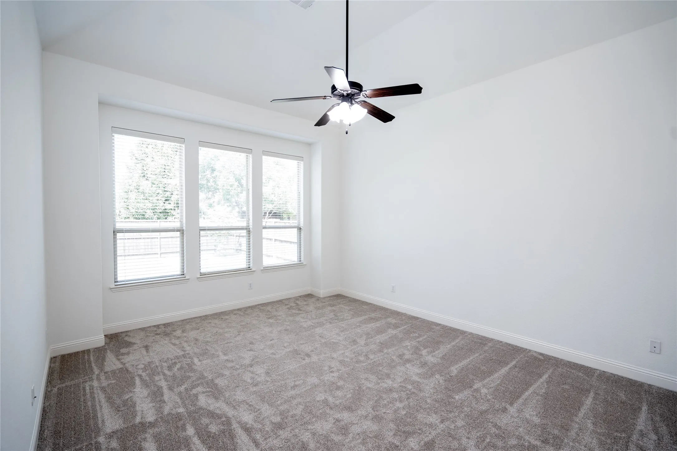 Vaulted ceiling, three large windows with blinds, ceiling fan with light fixture, neutral-toned carpet, and white walls
