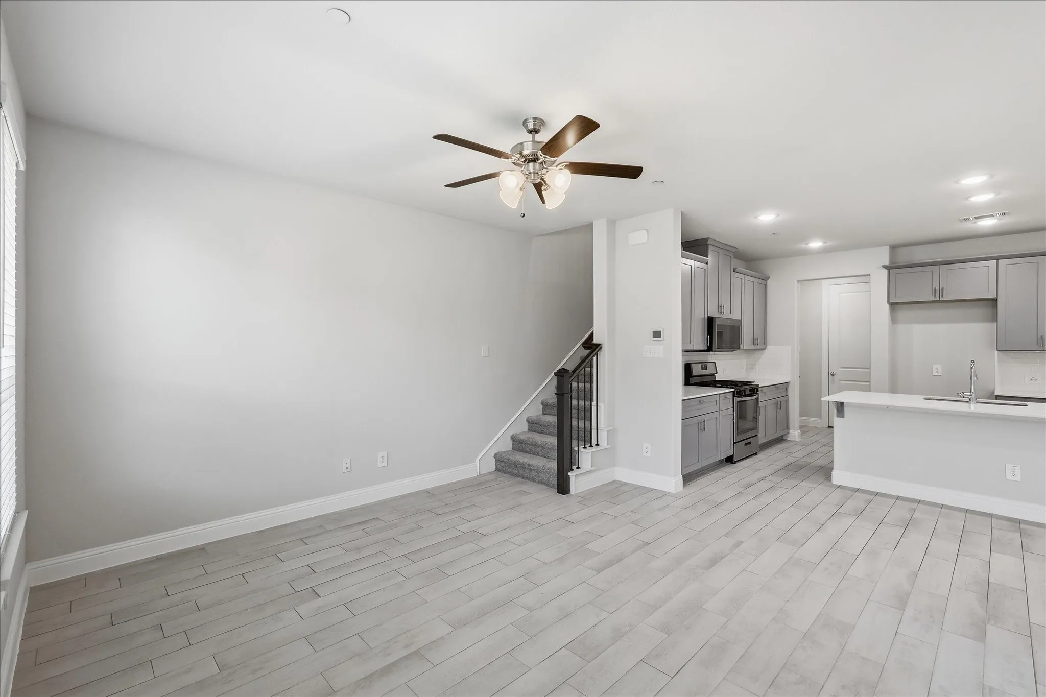 Open concept living area with wood-finish flooring, a ceiling fan, and a kitchen featuring stainless steel appliances, light gray cabinetry, and a peninsula with a sink