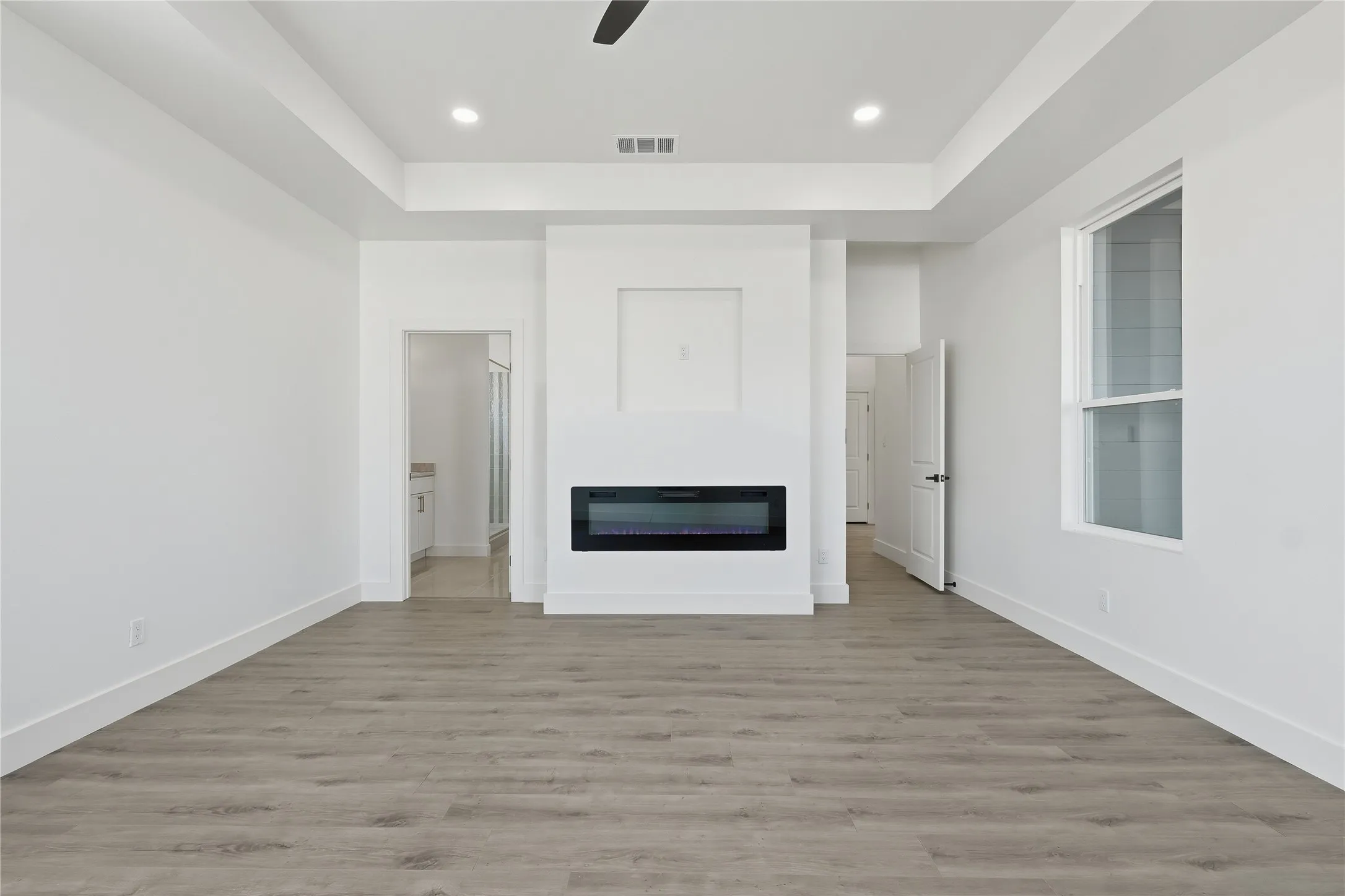 Spacious room featuring wood-finish flooring, a built-in electric fireplace, a tray ceiling with recessed lighting, and a window with a white frame