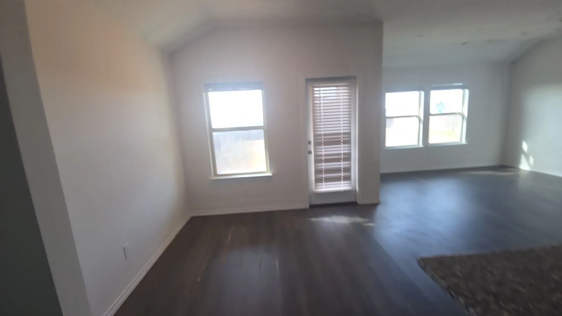 Vaulted ceiling, multiple windows with blinds, wood-finish flooring, and a glass-paneled door