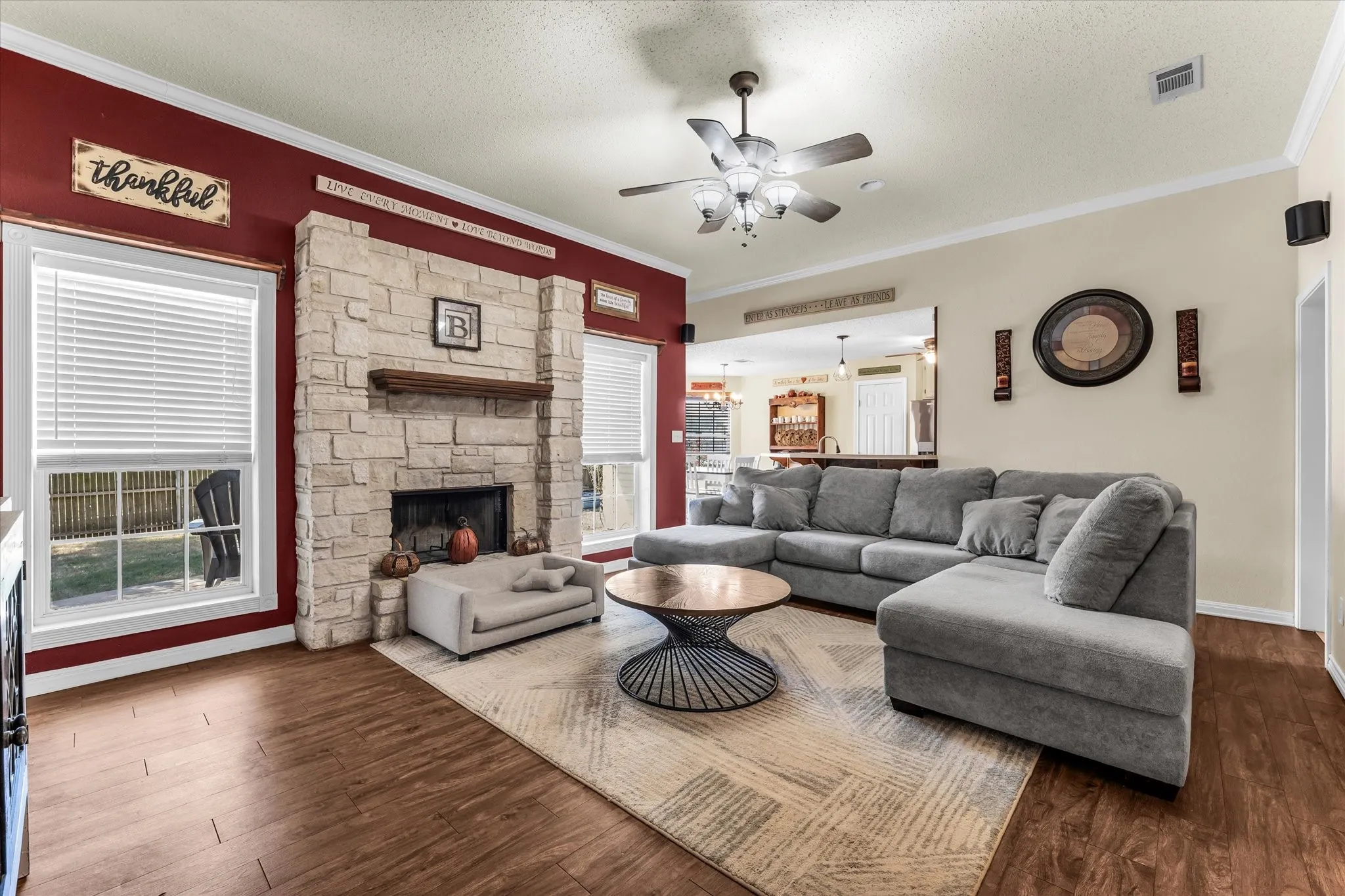 Stone-clad fireplace with a wood mantle, wood-finish flooring, two windows with blinds, ceiling fan, and crown molding