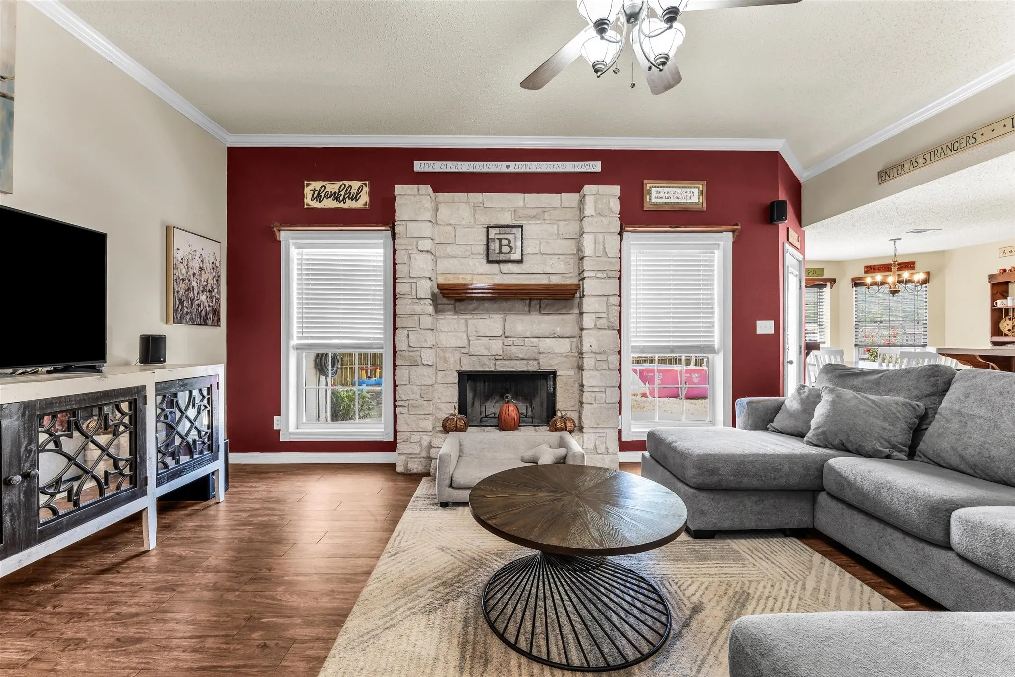 Stone fireplace with wood mantel, wood-finish flooring, crown molding, ceiling fan, and large windows with blinds