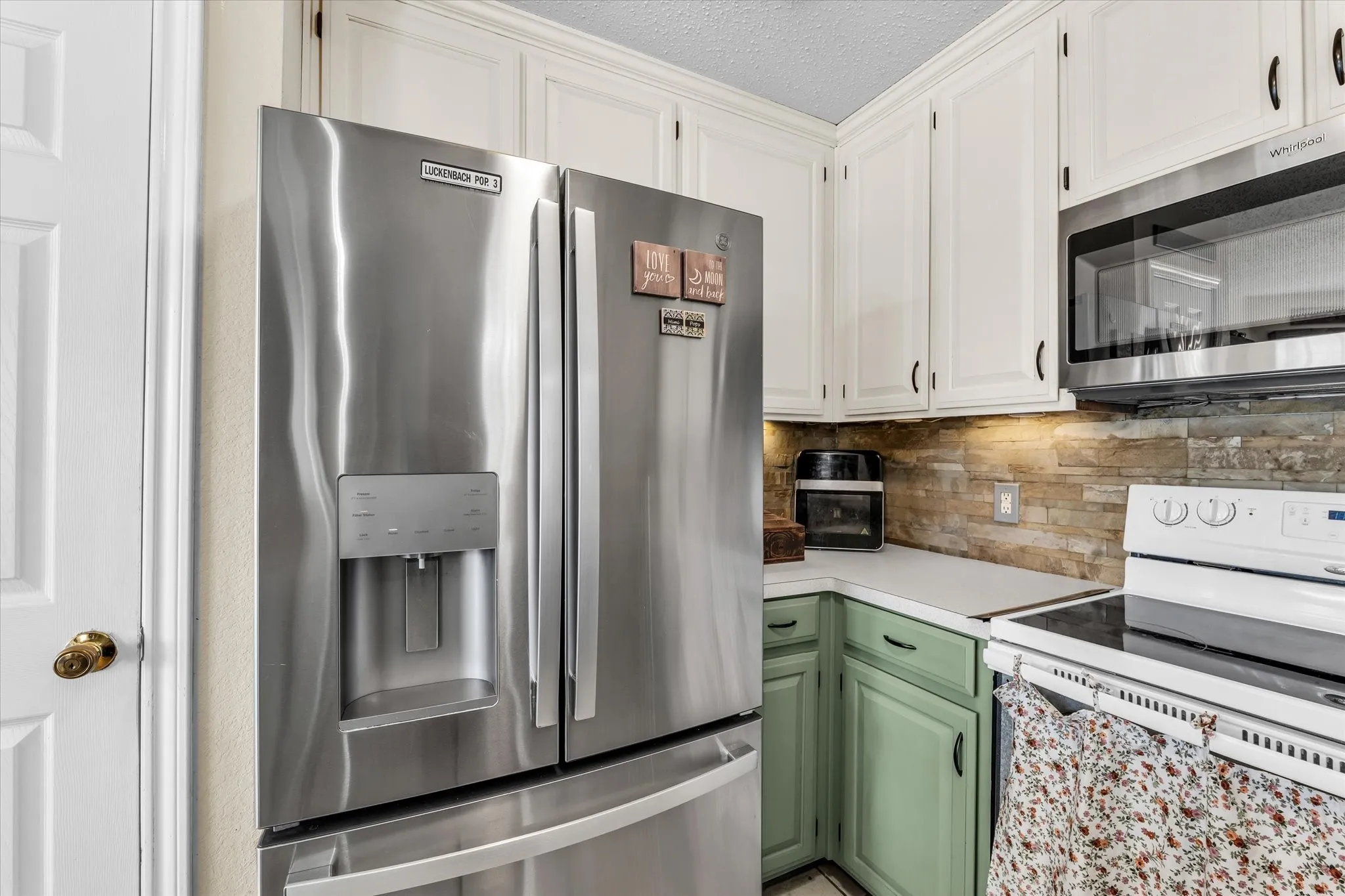 Stainless steel French door refrigerator with ice and water dispenser, white upper cabinetry, light green lower cabinetry, white countertops, and a stacked stone-look backsplash