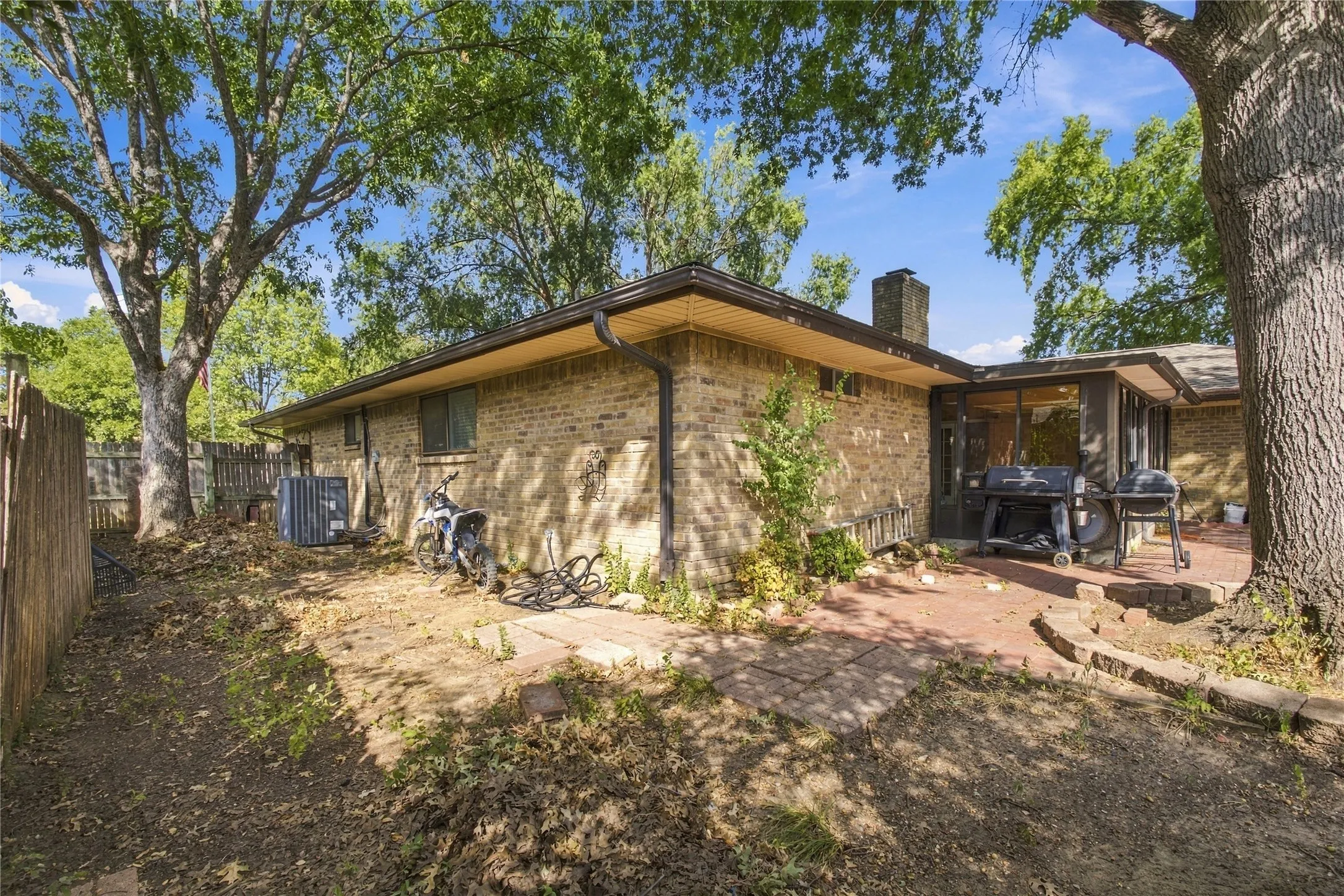 Brick exterior with a prominent chimney, large mature trees providing shade, and a brick paver patio
