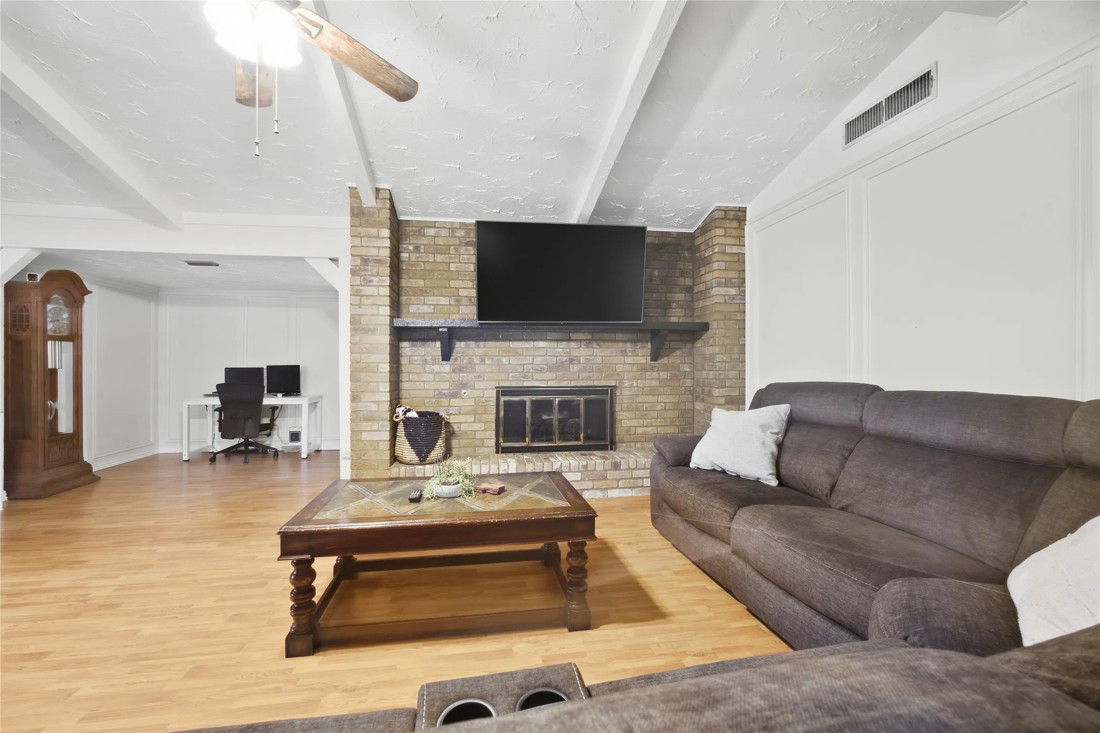 Wood-finish flooring throughout, brick fireplace with mantel, exposed ceiling beams, ceiling fan, and white paneling