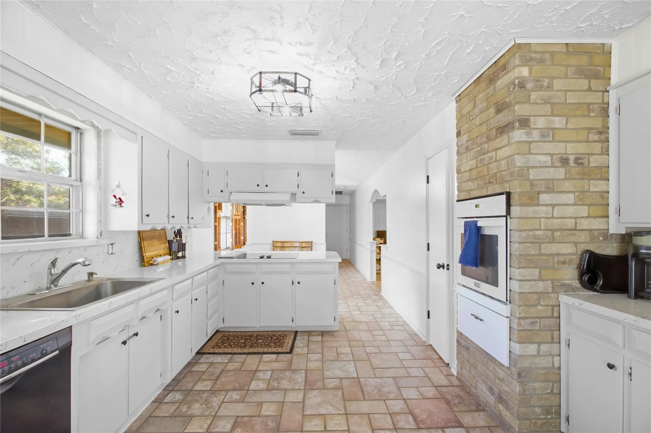 Kitchen featuring white cabinetry, light-toned brick accents, tile flooring, a built-in oven, and a double-basin stainless steel sink