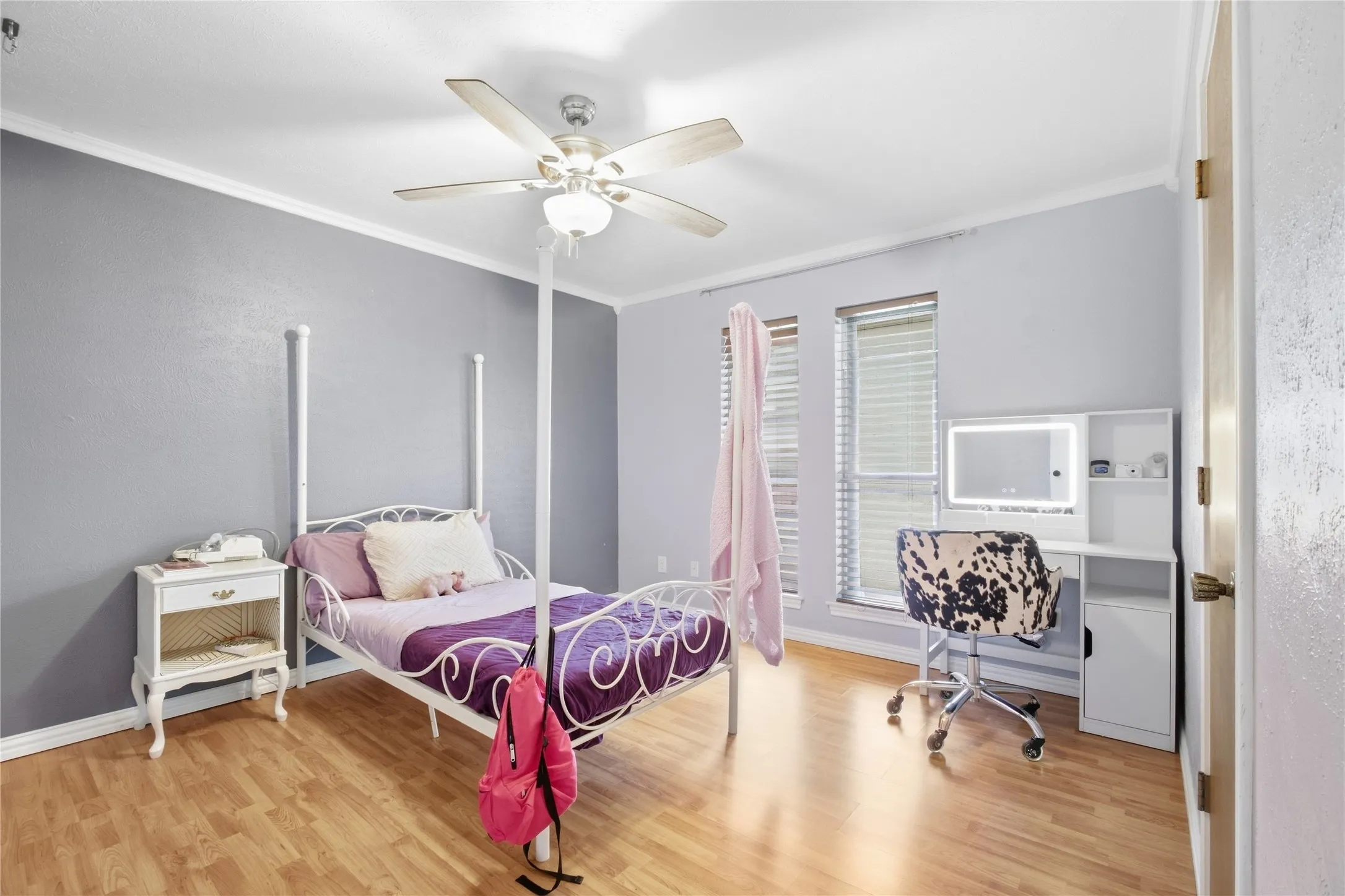 Wood-finish flooring, ceiling fan with integrated lighting, multiple windows with blinds, built-in desk with integrated lighting, and crown molding