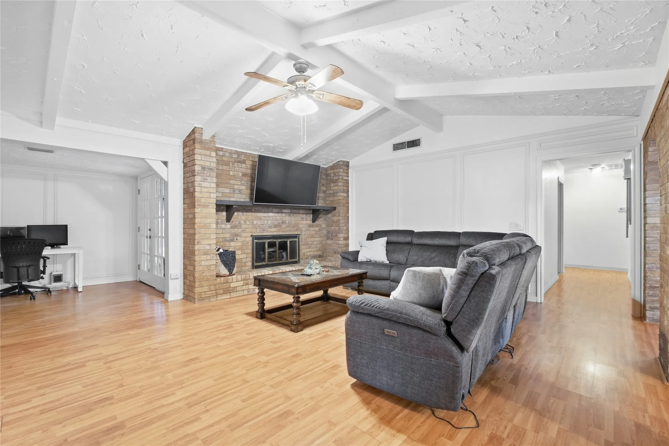 Spacious open-concept living area featuring coffered ceiling with ceiling fan, gleaming wood-finish flooring throughout, and a brick fireplace with mounted television
